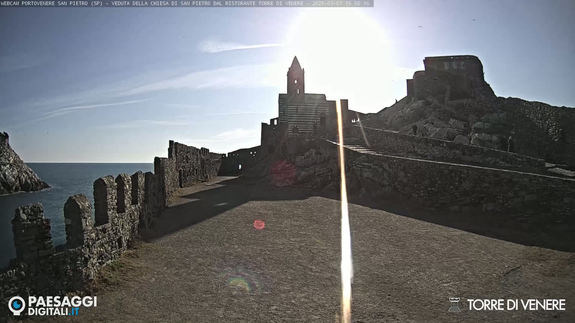 A stone pathway leads toward a striped church and castle ruins overlooking a calm ocean under a bright, sunny sky.