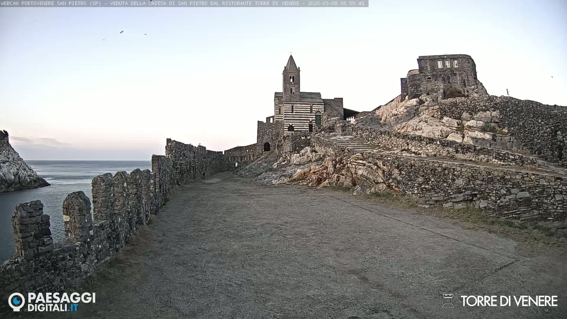 A stone pathway leads to a striped church and castle ruins on a rocky hill overlooking a calm sea under a clear sky.