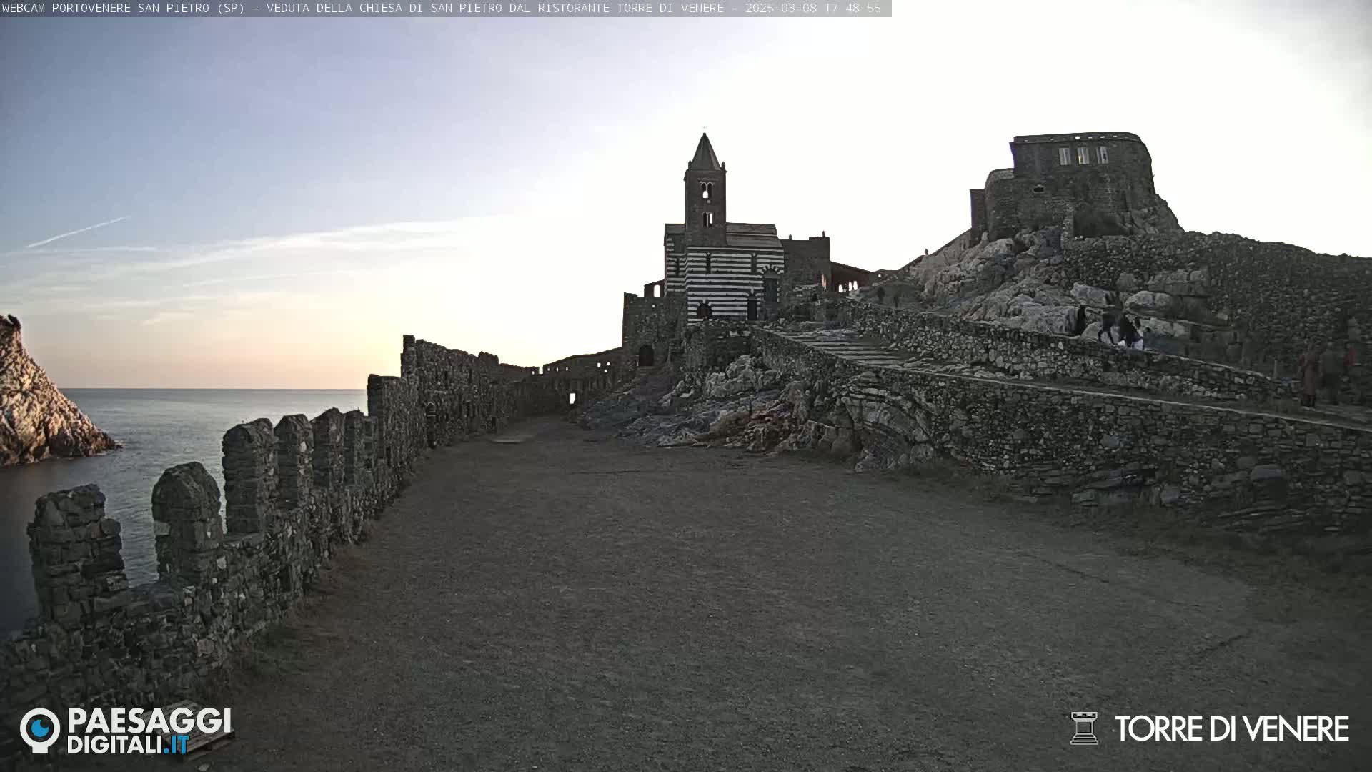 A stone church and castle ruins are situated on a rocky hill overlooking a calm sea under a clear, twilight sky.
