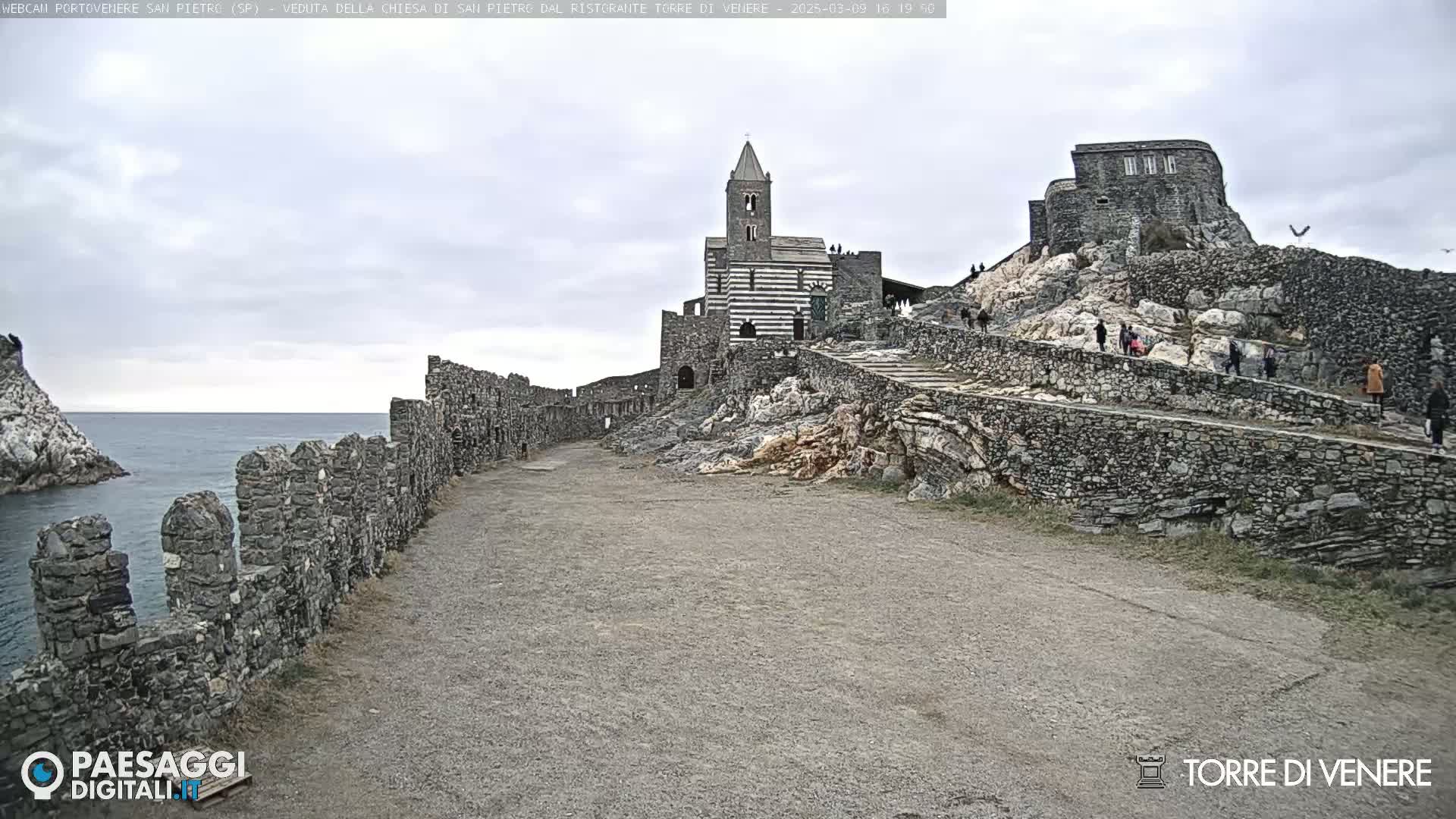A stone church and castle are situated atop a rocky hill overlooking a calm ocean under an overcast sky.