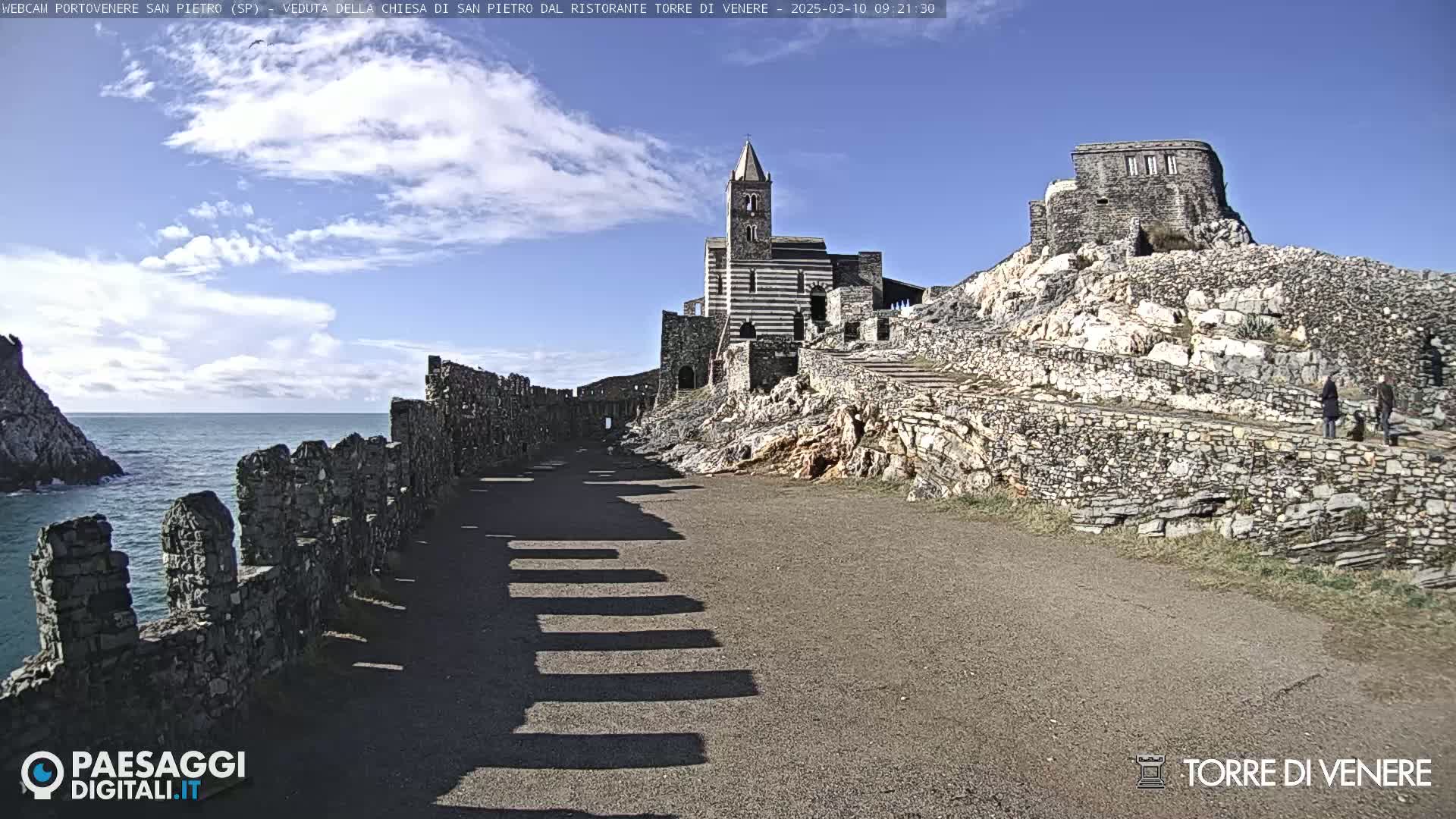 A stone path leads to a striped church on a rocky hill overlooking a calm ocean under a partly cloudy sky.