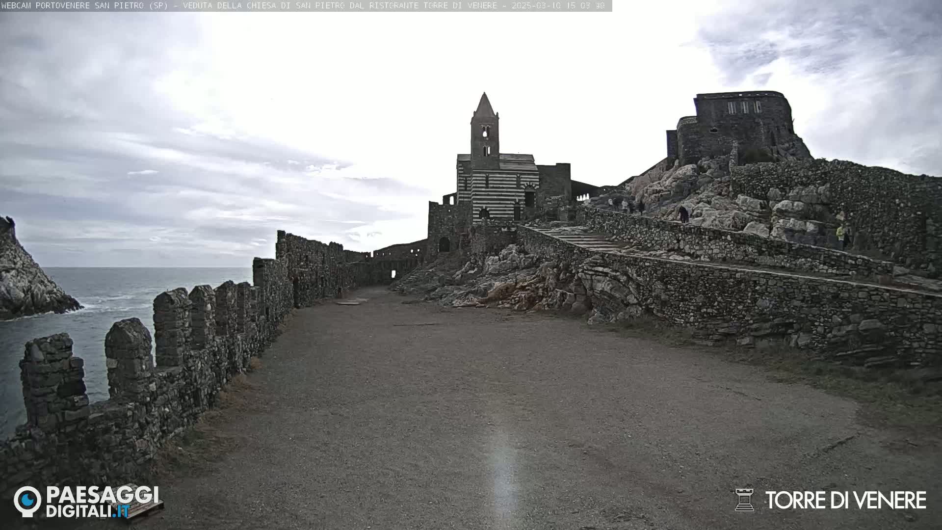 A stone pathway leads to a striped church and castle ruins overlooking a calm ocean under a cloudy sky.