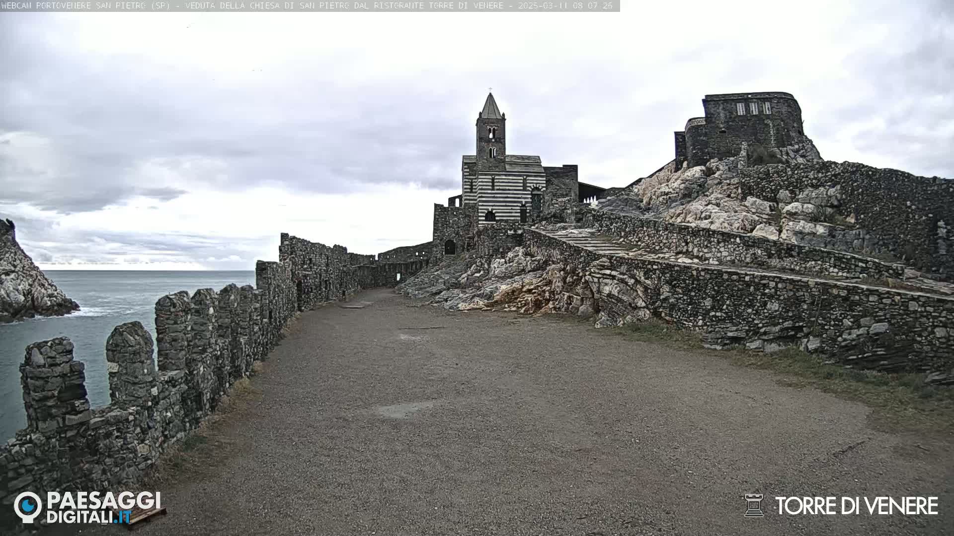 A stone pathway leads to a striped church and stone buildings atop a rocky cliff overlooking a calm sea under an overcast sky.