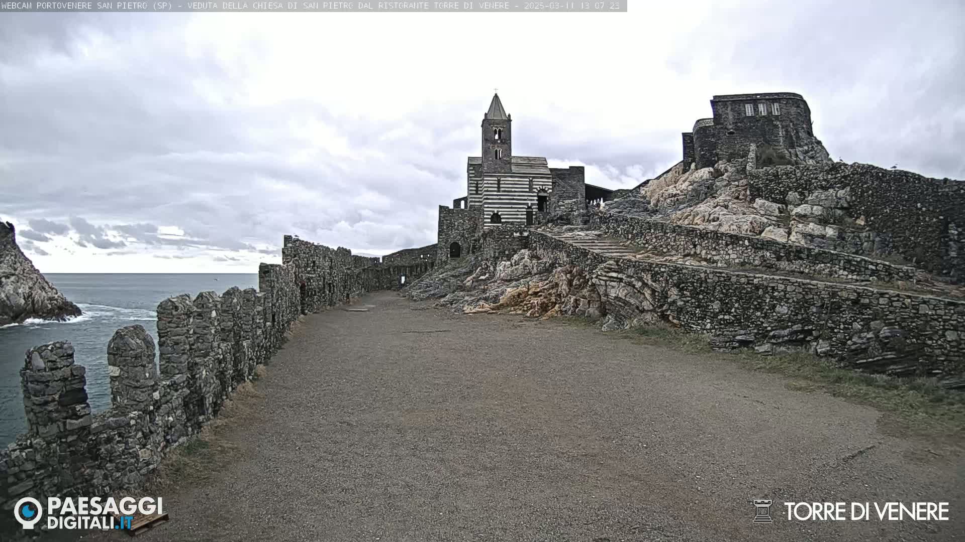 A stone pathway leads toward a striped church and stone ruins perched on a rocky cliff overlooking a calm ocean under a cloudy sky.