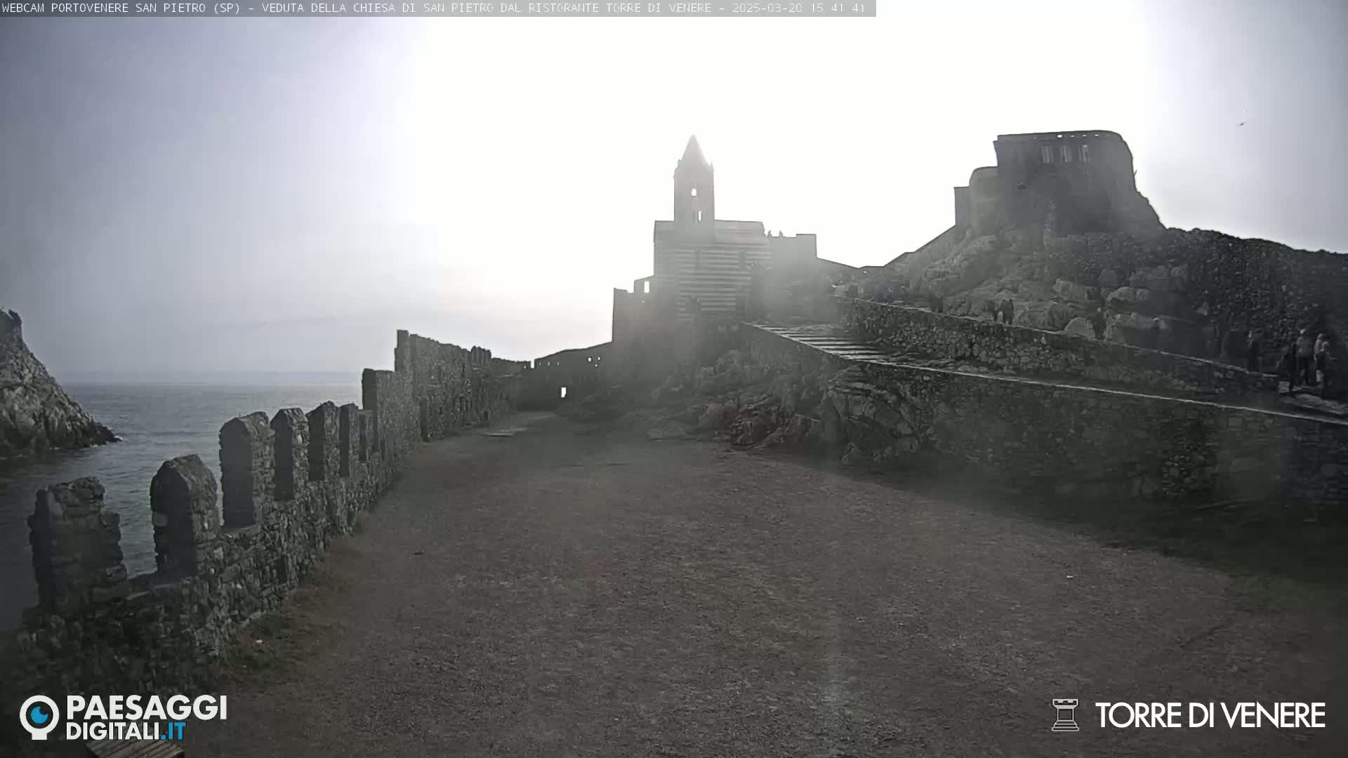 A hazy view of a stone church and fortress overlooking a calm sea, with a stone wall extending from the foreground to the water's edge.