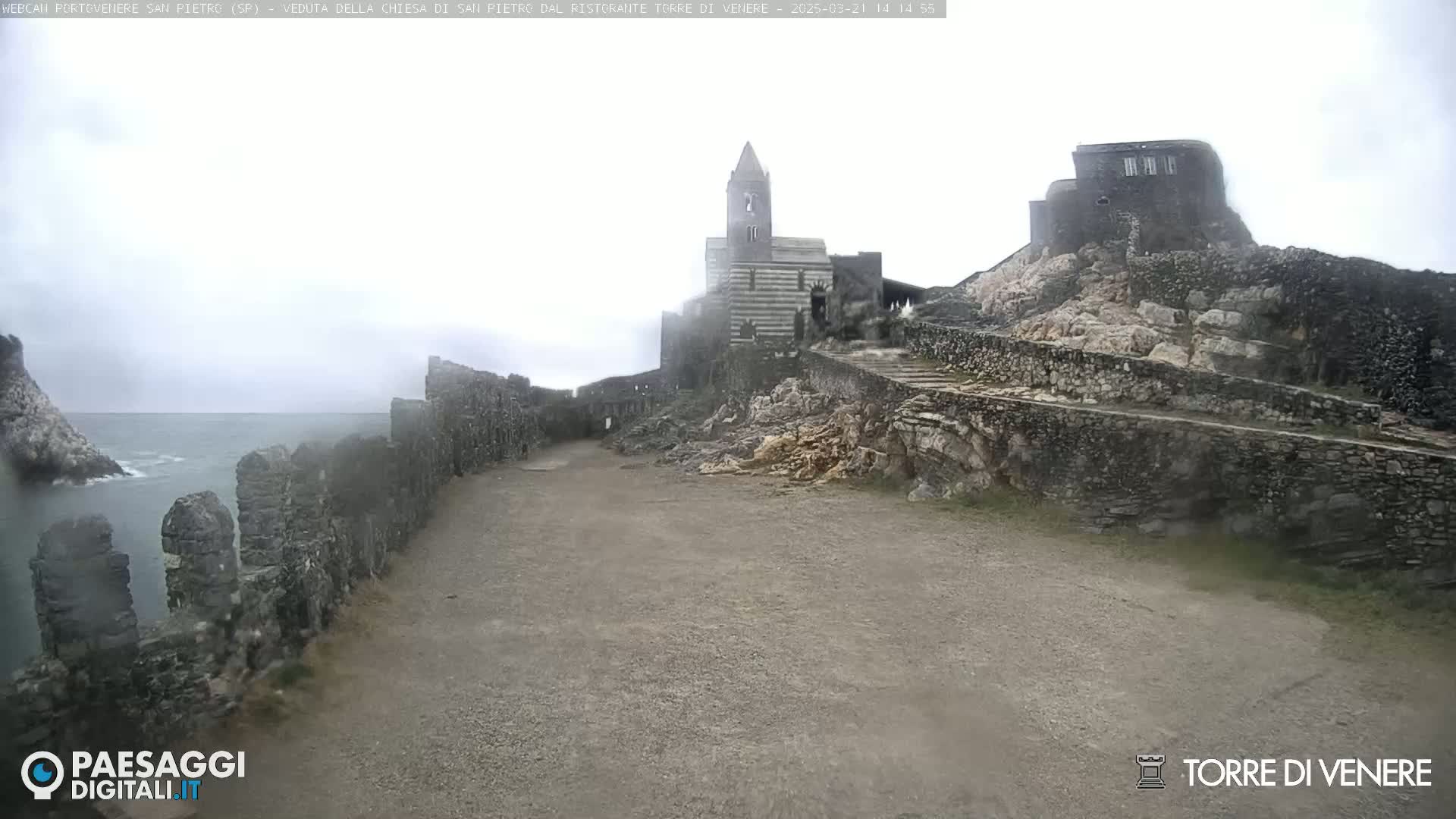 A stone church and fortress are situated on a rocky hillside overlooking a calm sea under an overcast sky.
