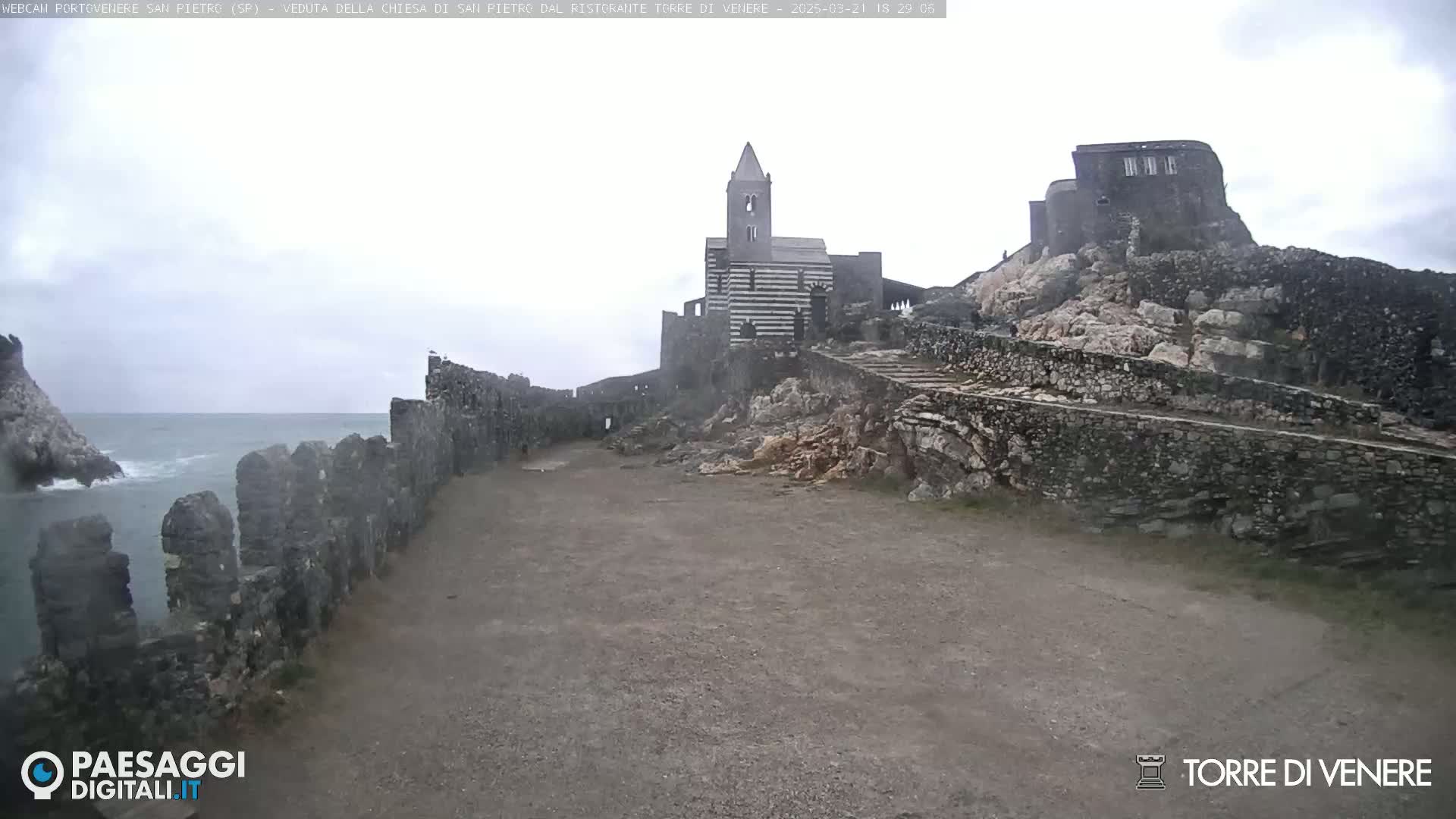 A stone pathway leads to a striped church and castle ruins overlooking a rough sea under an overcast sky.