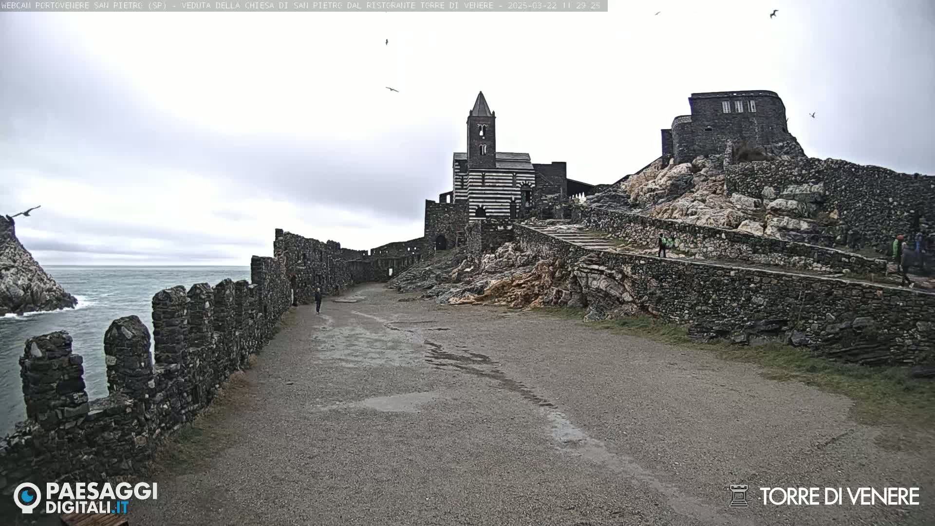 A stone pathway leads to a striped stone church situated on a rocky cliff overlooking a calm sea under an overcast sky.