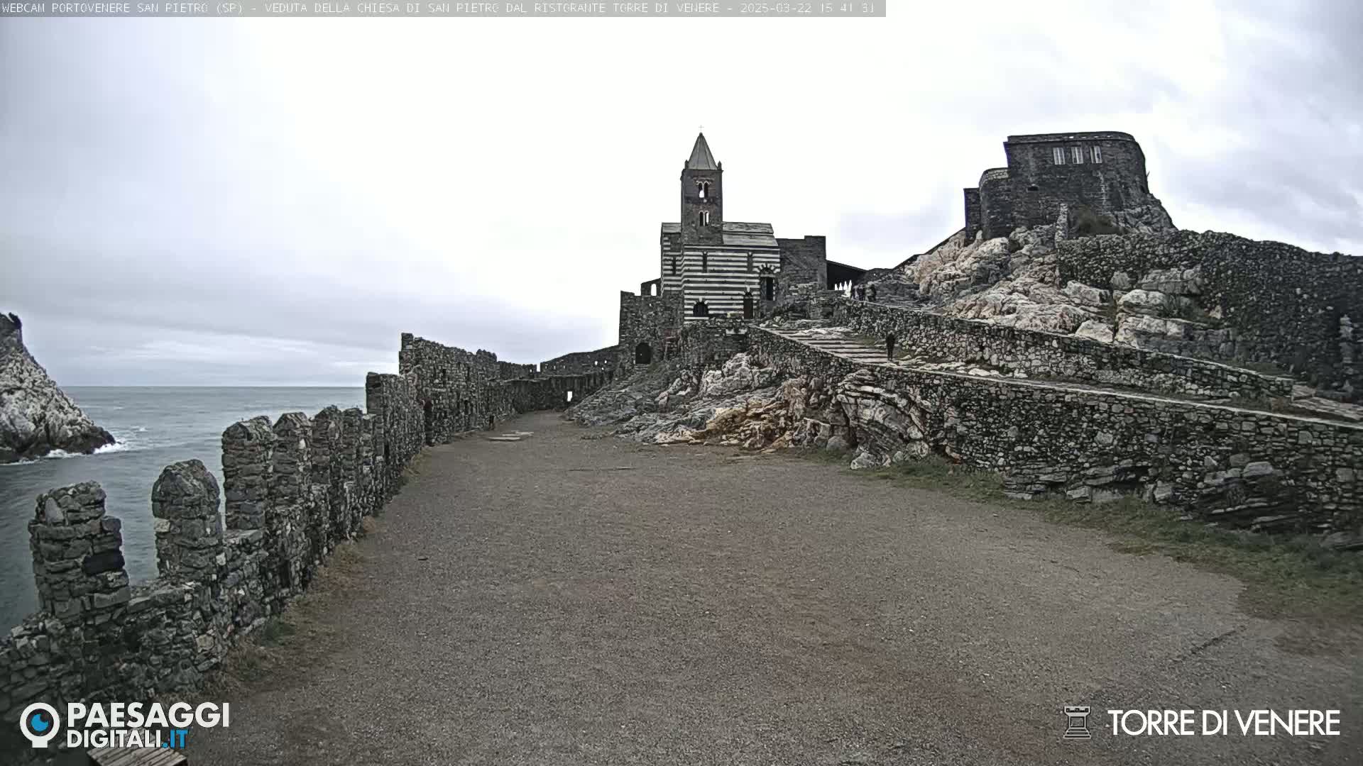A stone pathway leads to a striped church and castle ruins situated on a rocky cliff overlooking a calm sea under an overcast sky.