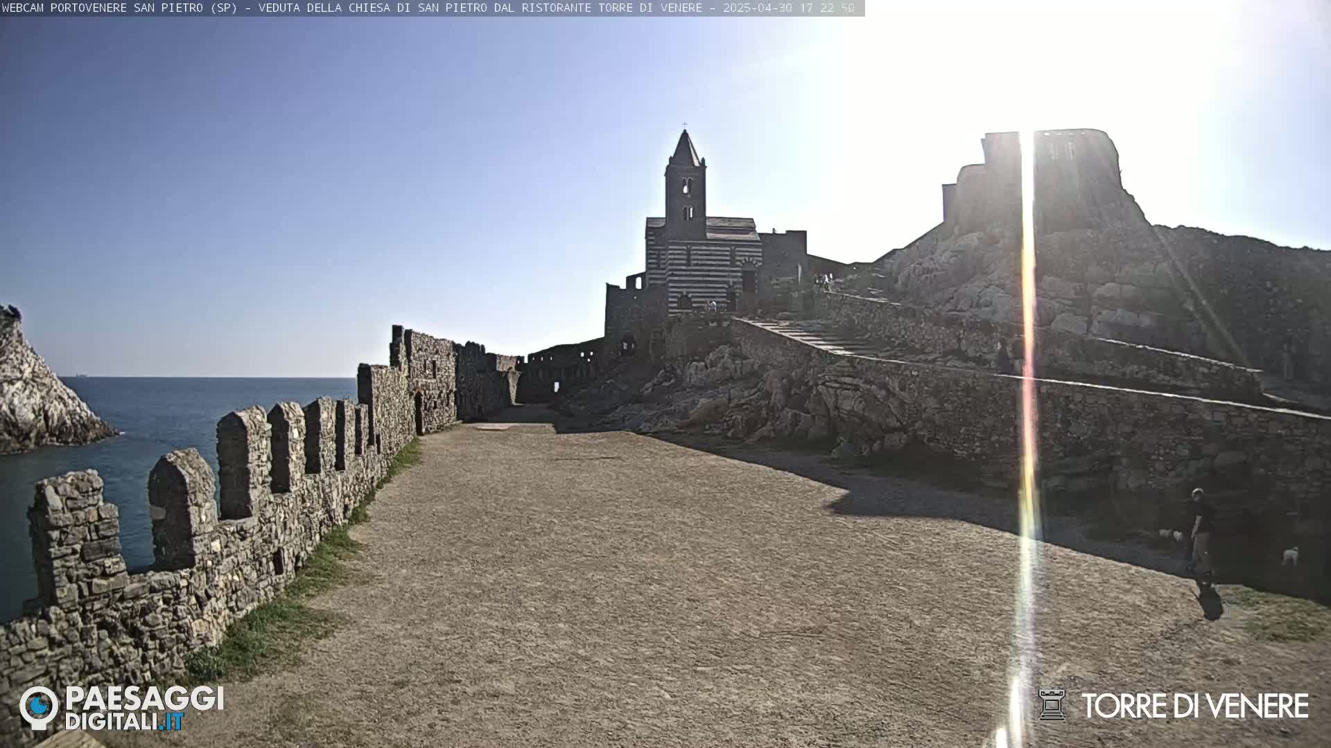 A sunny day reveals a coastal stone pathway leading to a striped church and castle ruins overlooking the ocean.