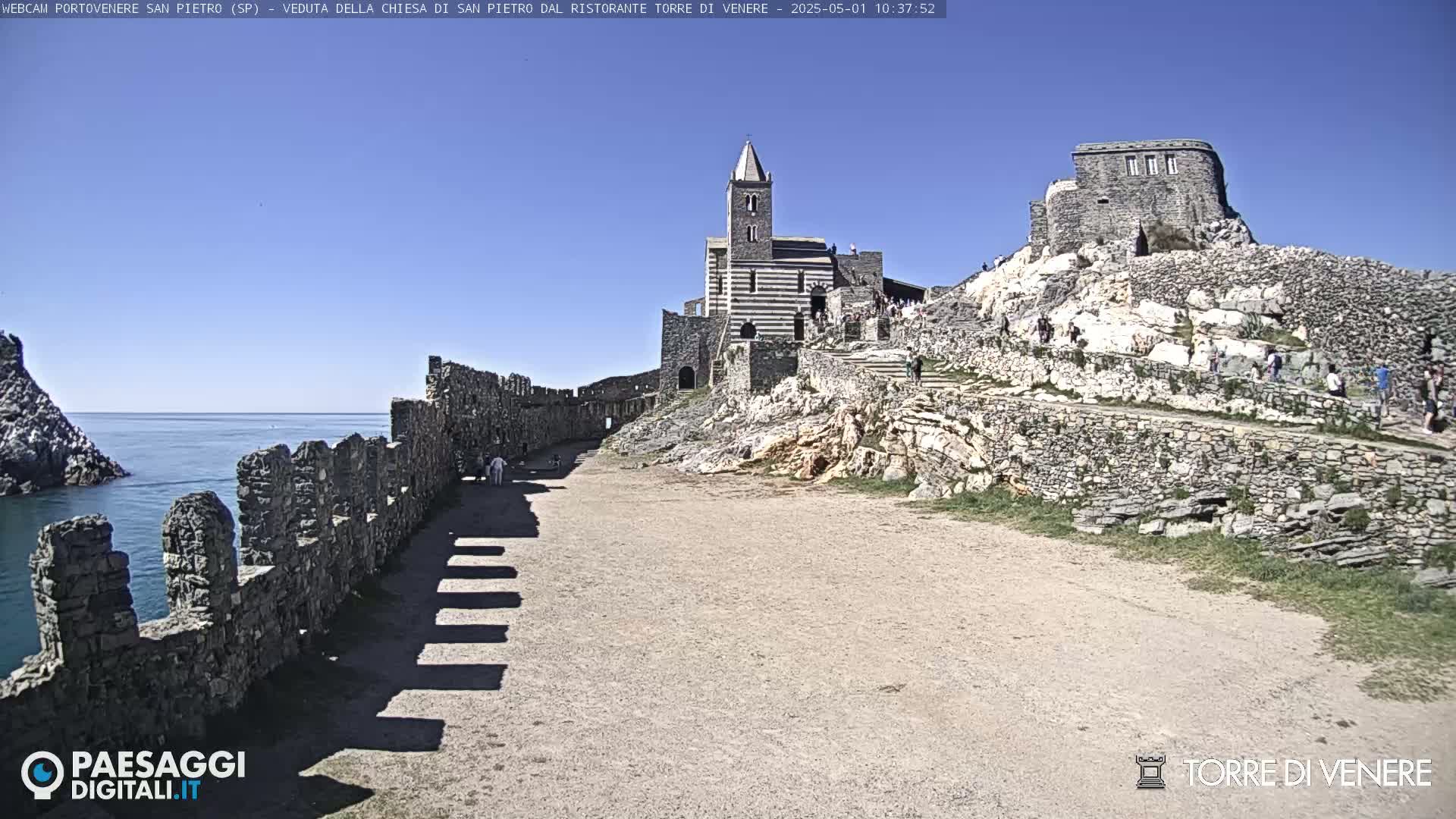 A stone church and castle ruins sit atop a rocky hill overlooking a calm ocean under a clear blue sky.
