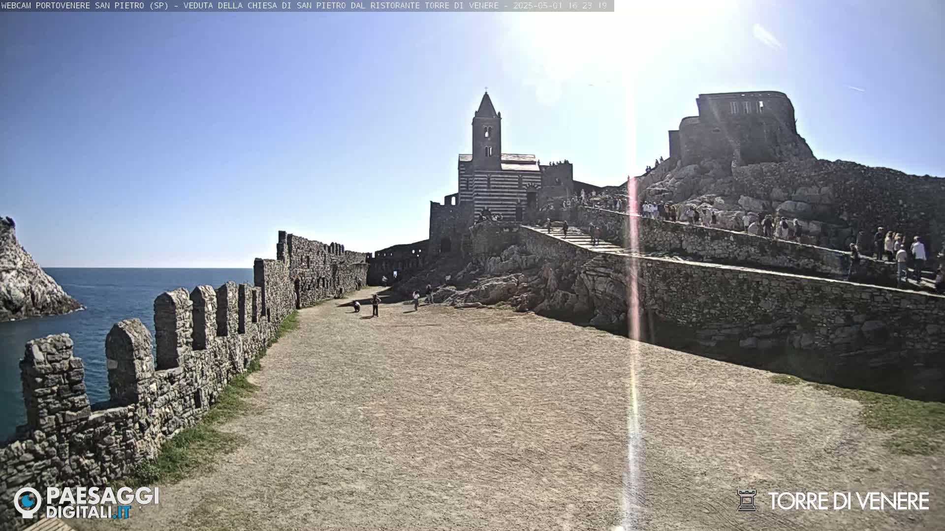 A stone church and castle ruins on a rocky hill overlooking a calm sea under a sunny sky are filled with many people.