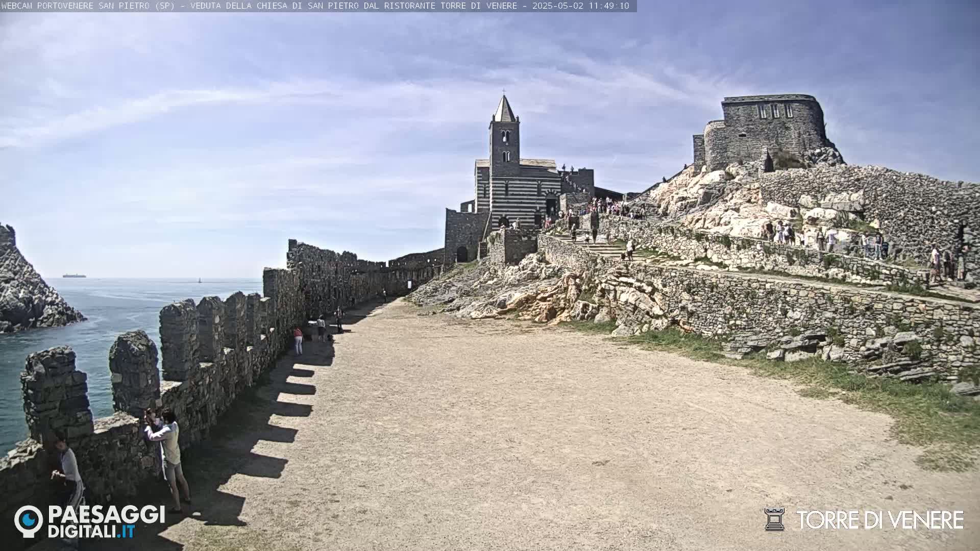 A coastal scene under a mostly clear sky shows a stone path leading to a striped church and castle ruins on a rocky hill overlooking the ocean.