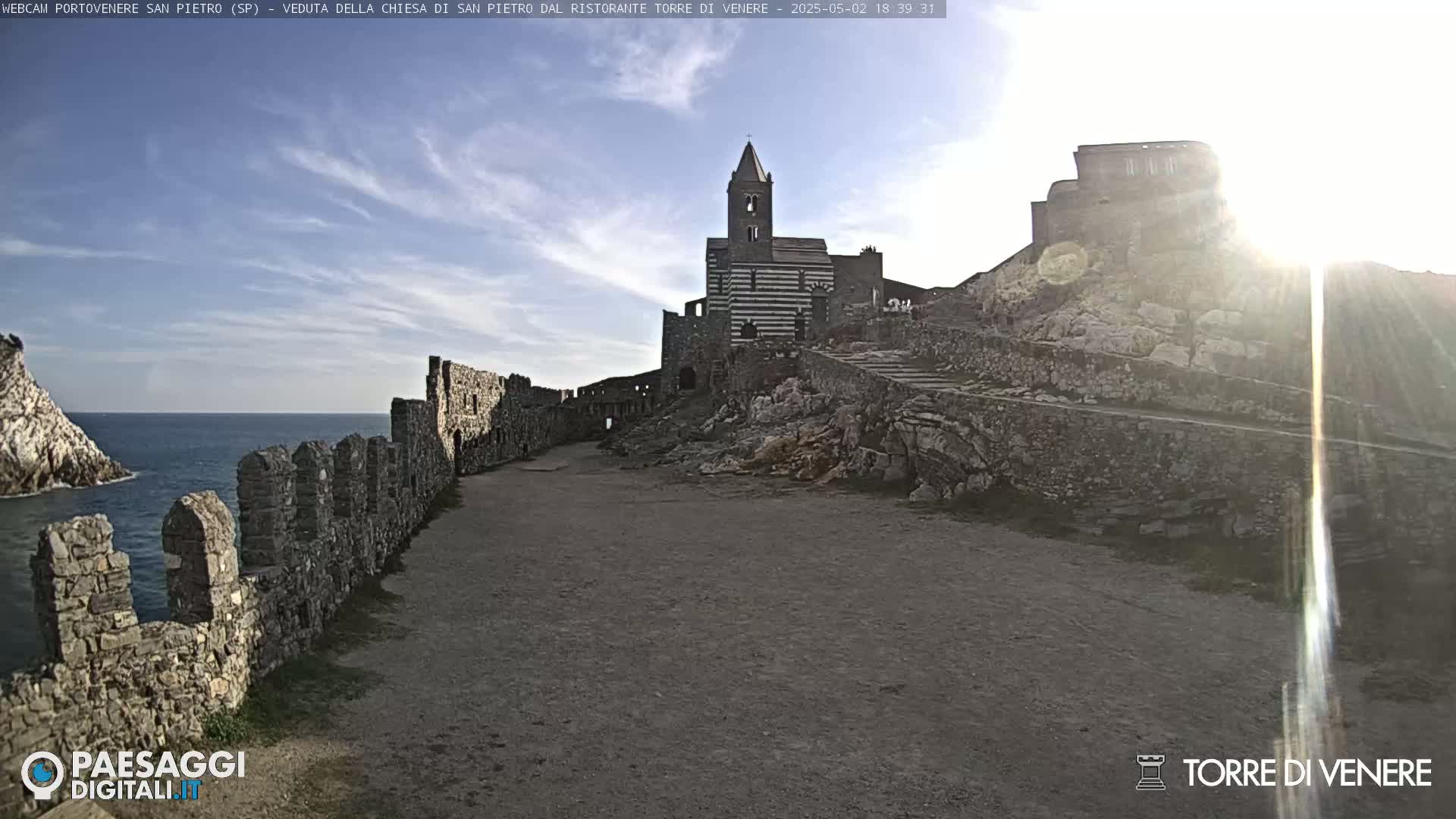 A stone pathway leads to a striped church and castle ruins overlooking a calm sea under a mostly sunny sky.