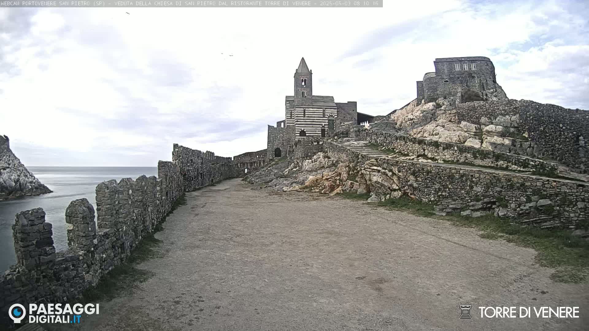 A stone path leads to a striped church on a rocky hill overlooking a calm sea under a cloudy sky.