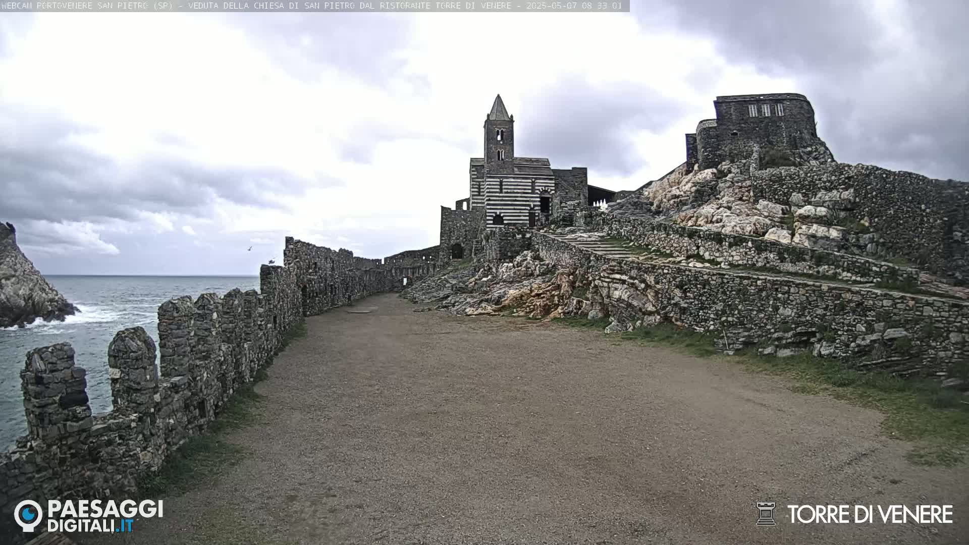 A stone path leads to a striped church and ruins on a rocky hill overlooking a wavy ocean under a cloudy sky.