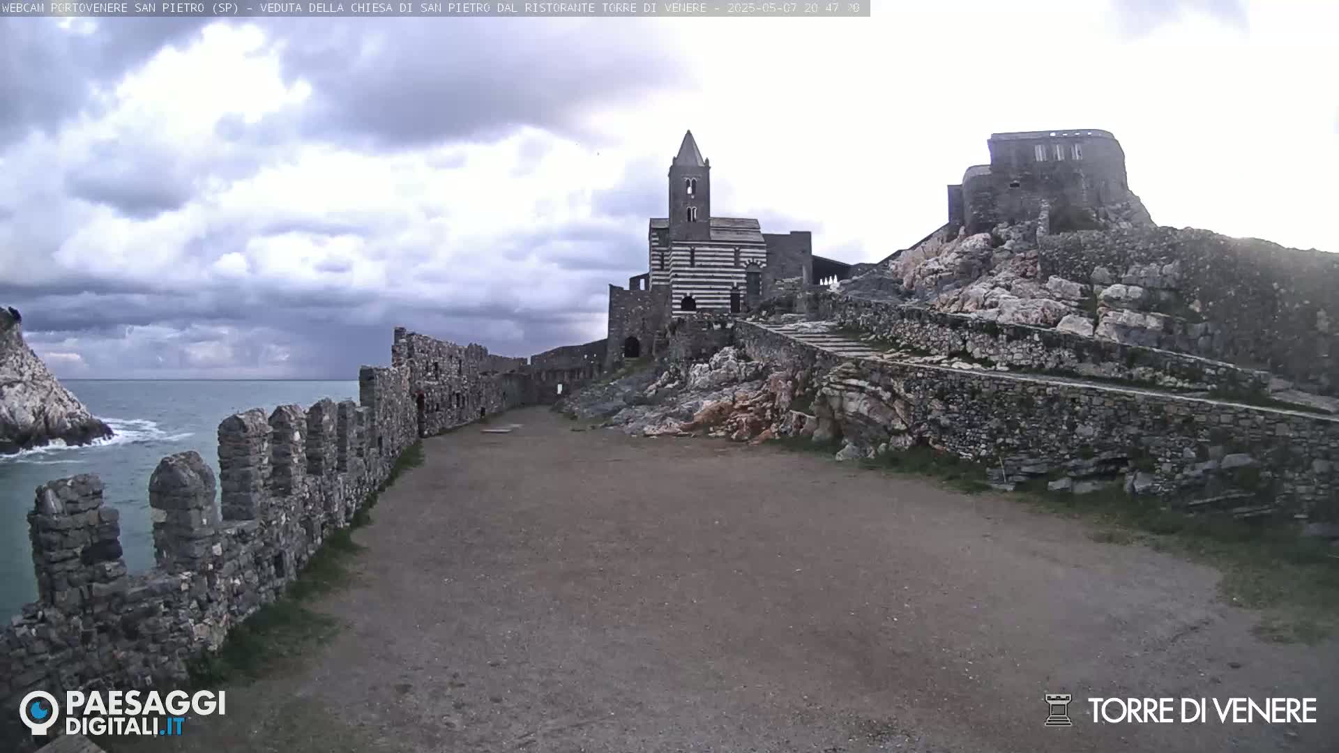 A stone pathway leads toward a striped stone church and castle ruins perched on a rocky hill overlooking a turbulent sea under a cloudy sky.