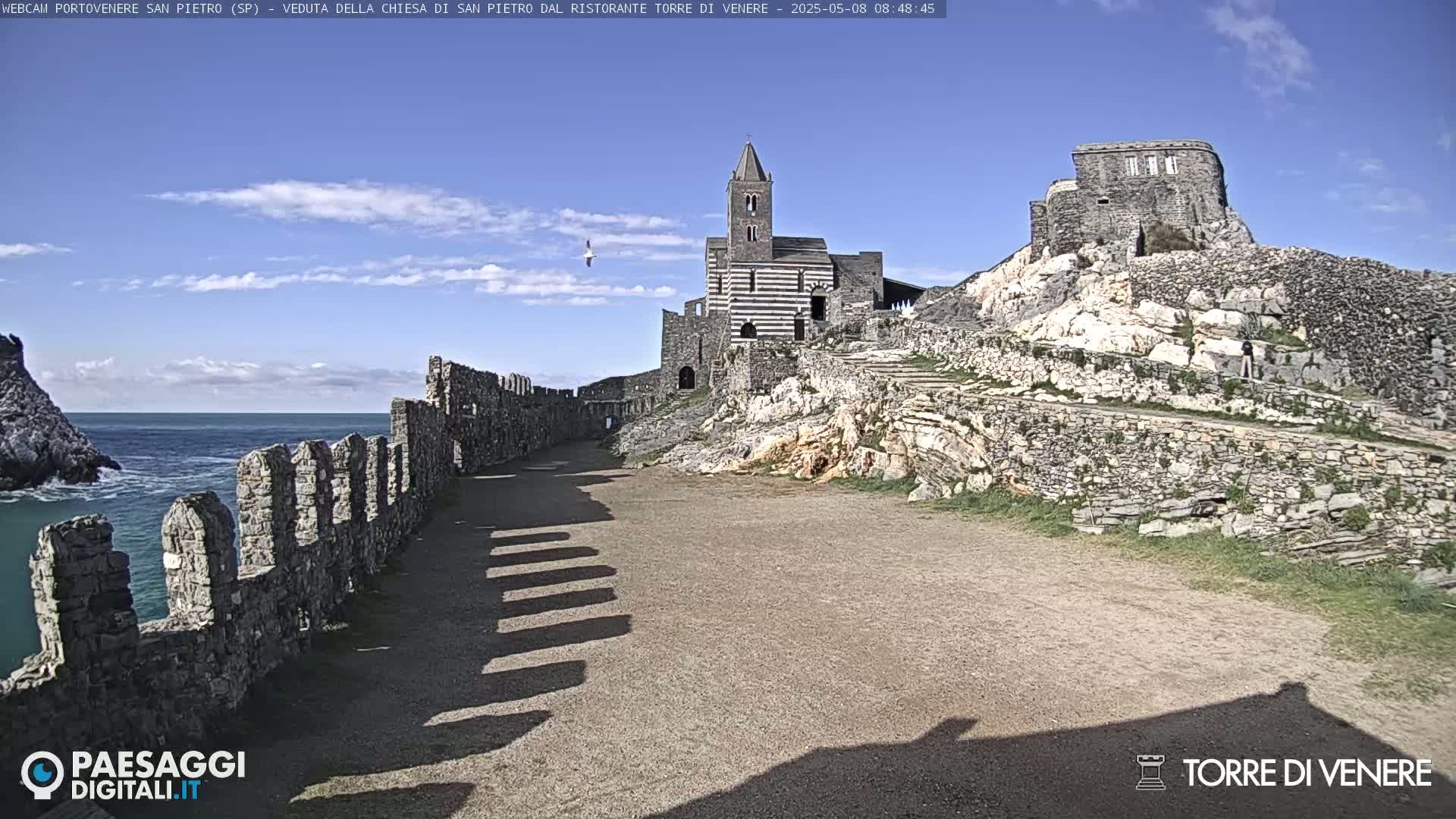 A stone pathway, lined by a low wall, leads toward a striped church and ruined castle perched on a rocky cliff overlooking a calm sea under a mostly clear sky.