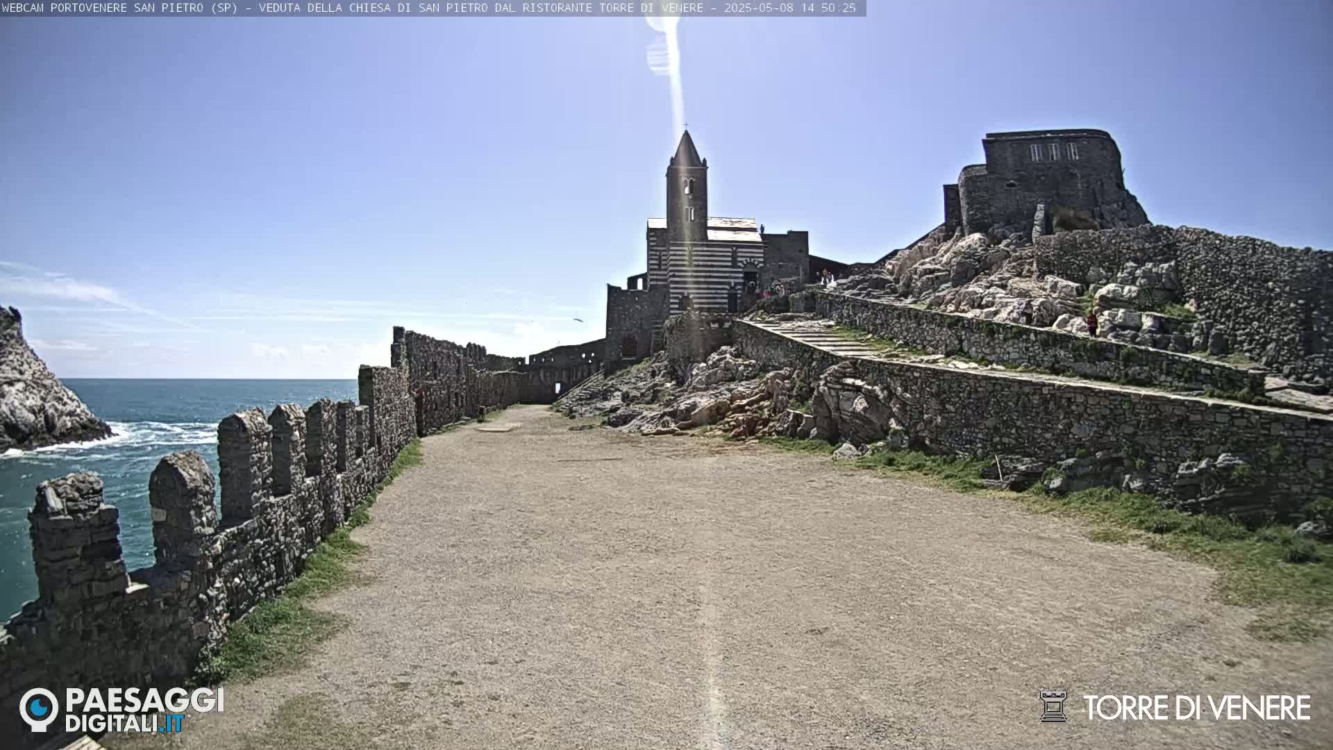 A stone path leads toward a striped church and stone ruins on a rocky hill overlooking a calm ocean under a clear blue sky.