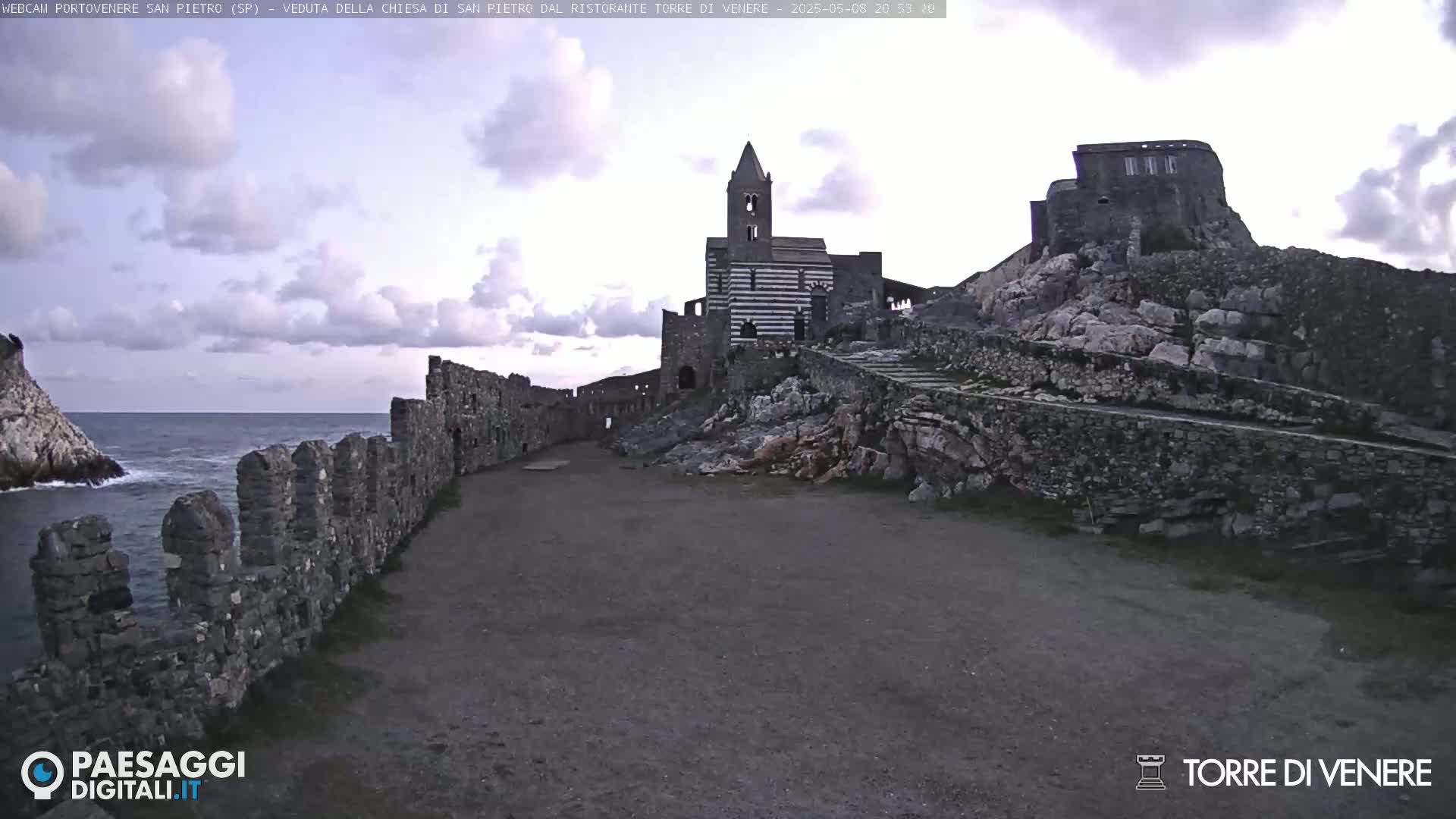 A stone path leads to a striped stone church and castle ruins overlooking a calm sea under a cloudy sky.