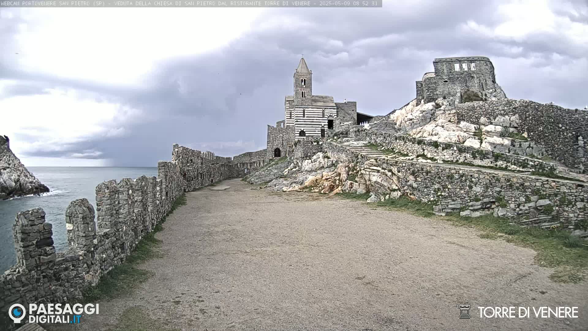 A stone church and ruins sit atop a rocky cliff overlooking a calm sea under a cloudy sky.