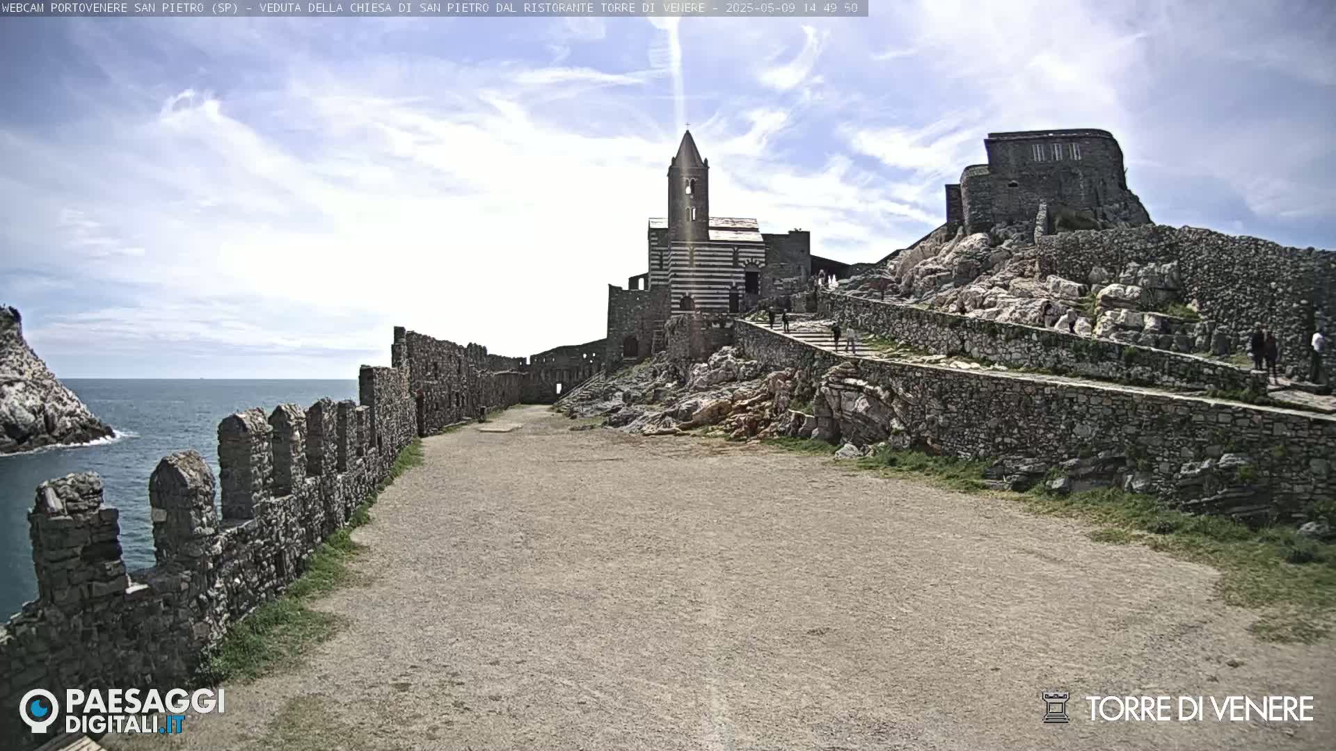 A stone path leads to a striped church atop a rocky hill overlooking a calm sea, under a partly cloudy sky.