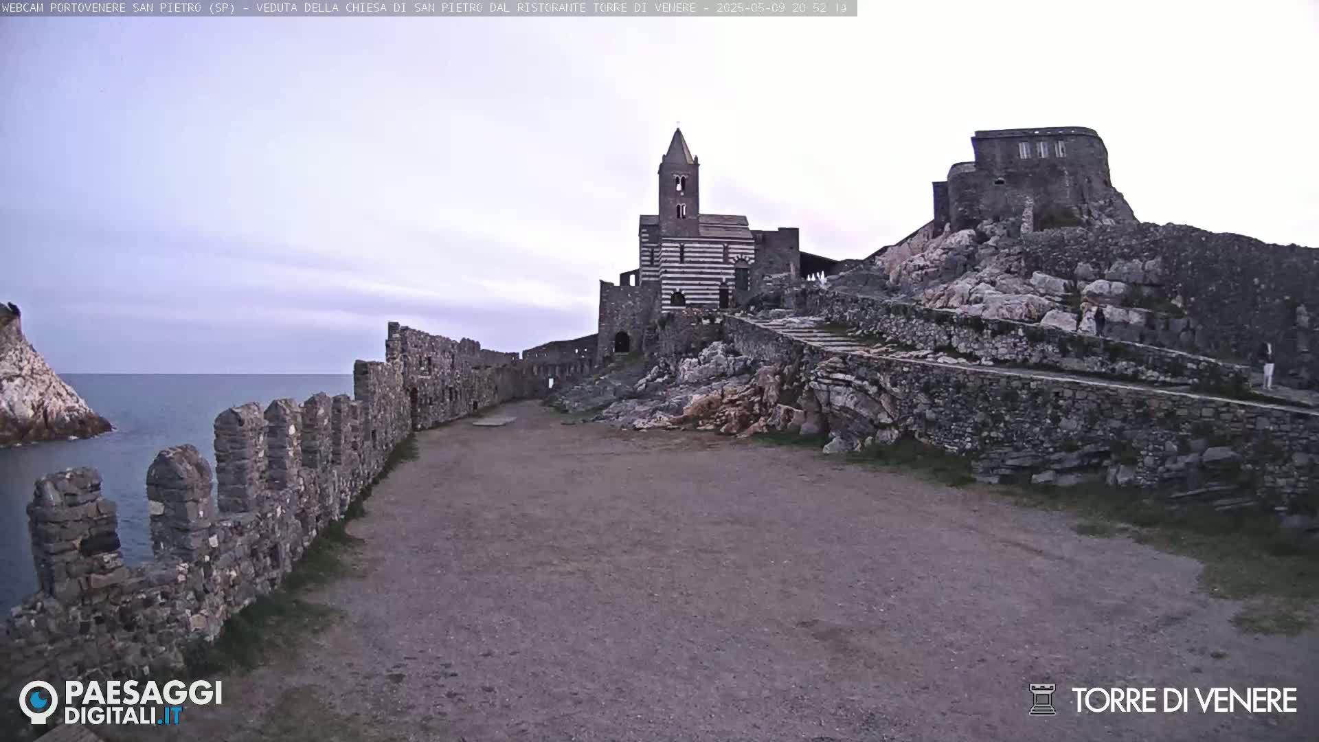 A stone pathway leads to a striped church and castle ruins overlooking a calm sea under a mostly clear sky.