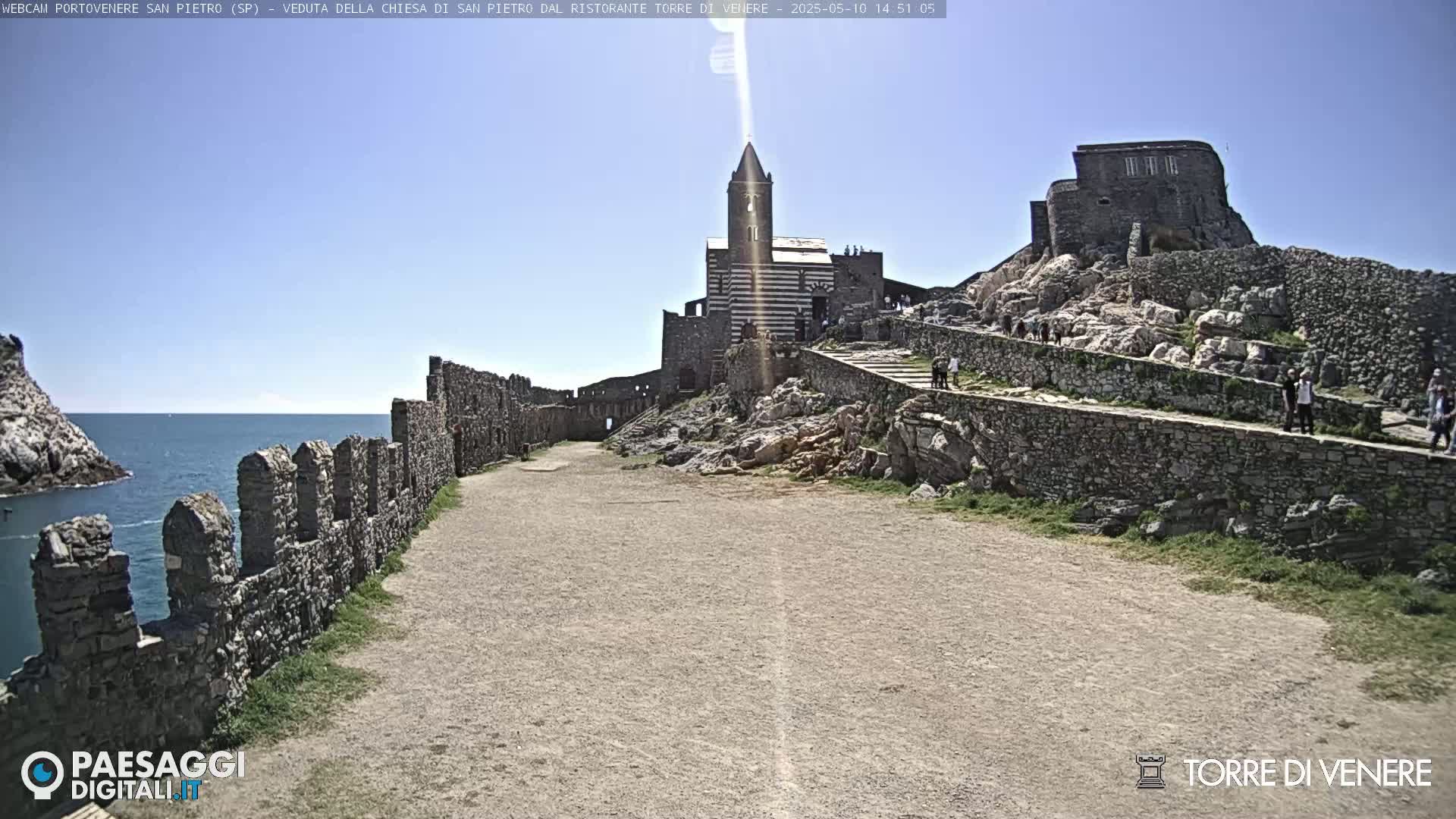 A coastal path leads to a striped church and castle ruins on a sunny day, with a calm sea visible in the background.