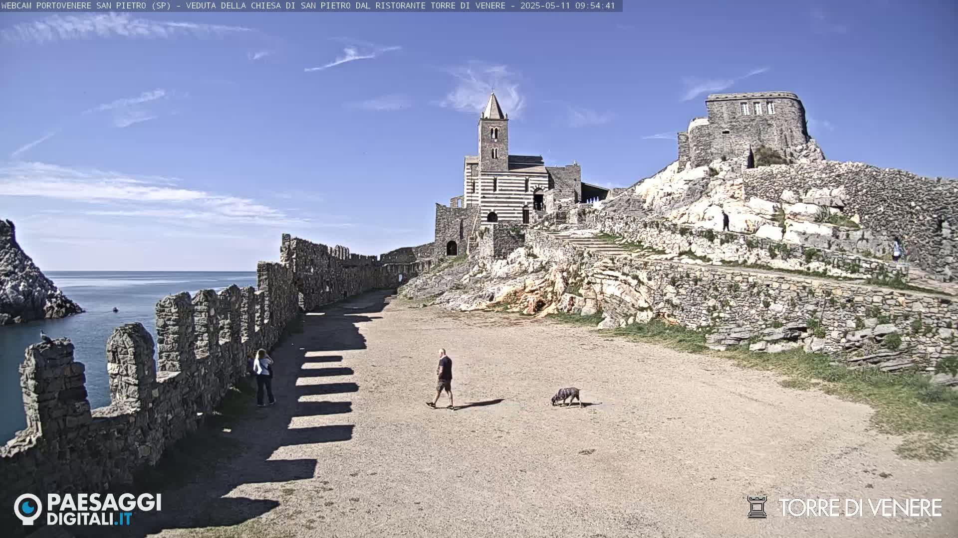 A coastal scene under sunny skies shows a stone pathway between ancient ruins leading to a striped church, with a man and his dog walking along the path.