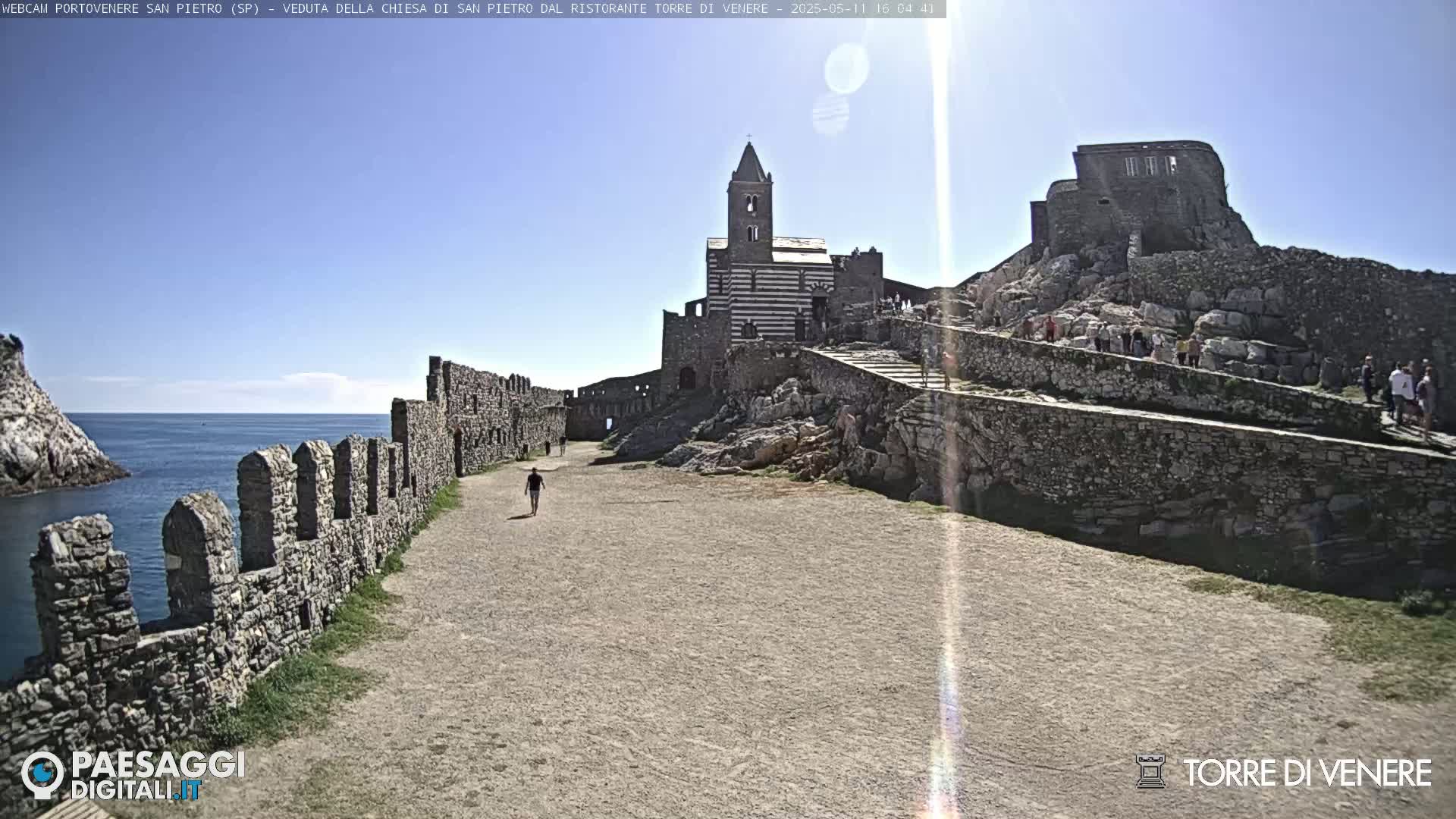 A stone pathway leads toward a striped church and castle ruins perched on a rocky hillside overlooking a calm sea under a sunny sky.