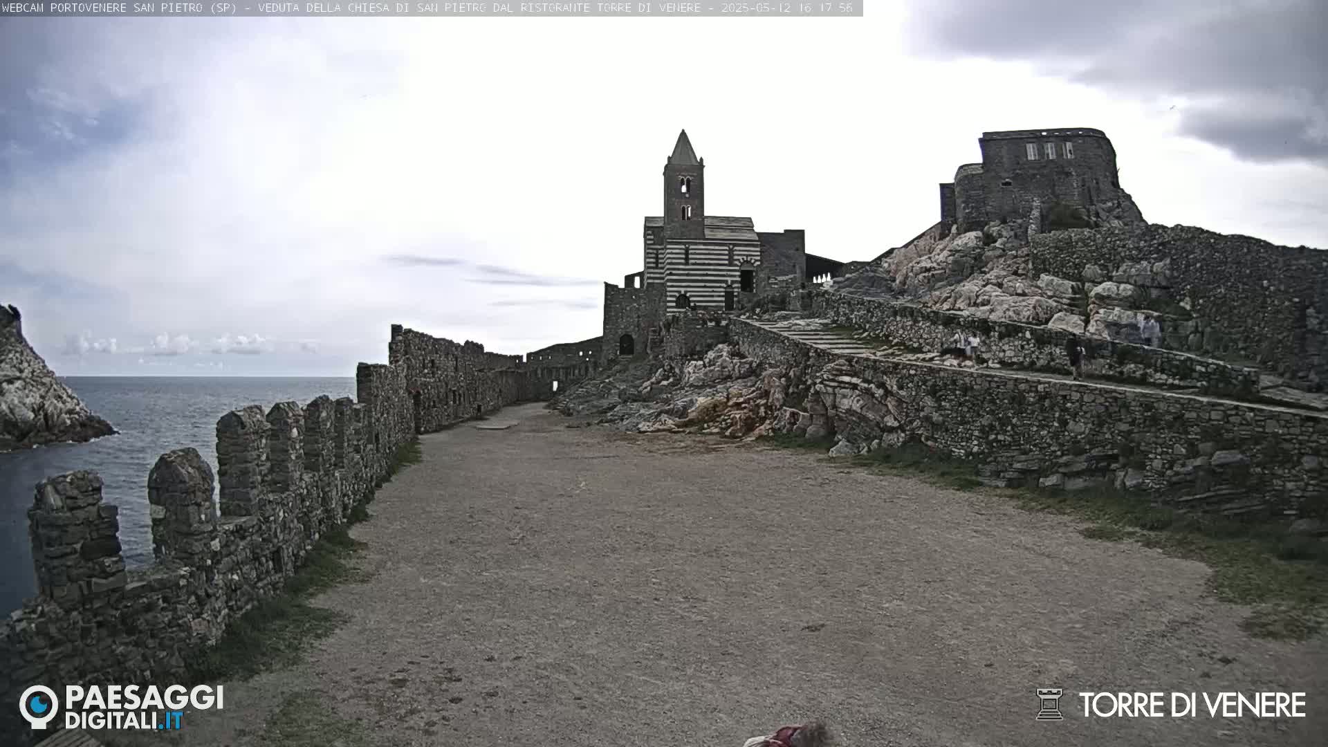 A stone pathway leads to a striped stone church and castle ruins situated on a rocky cliff overlooking a calm sea under an overcast sky.