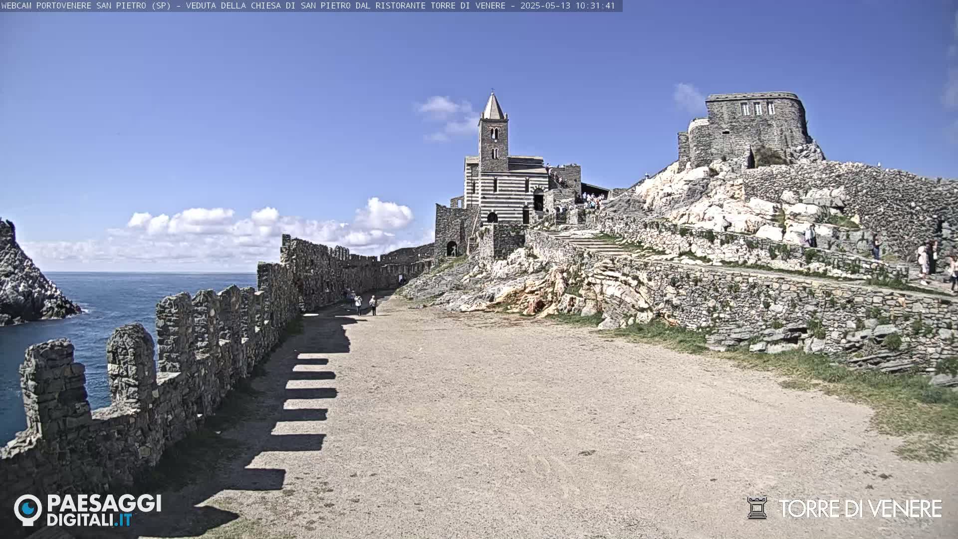 A stone pathway leads to a striped church and ruins on a rocky hill overlooking a calm ocean under a mostly sunny sky.