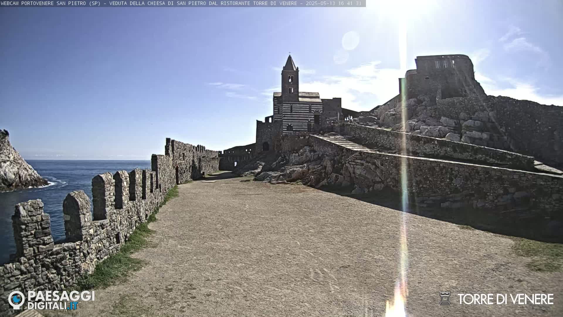 A stone church and castle ruins stand on a rocky outcrop overlooking a calm sea under a sunny sky.