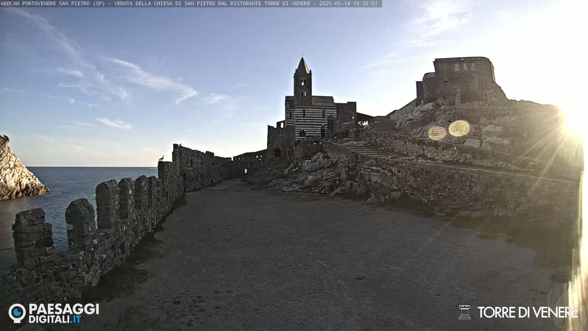 A stone path leads to a striped church and ruins perched on a rocky cliff overlooking a calm sea under a mostly clear sky.