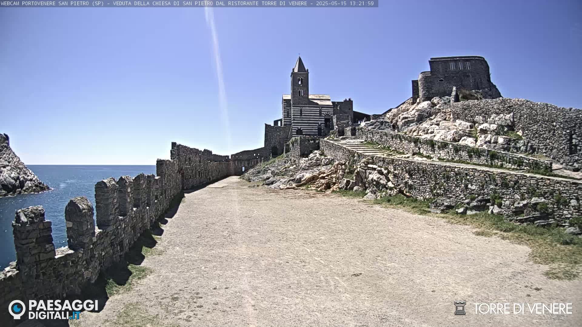 A stone pathway leads to a striped church and castle ruins on a sunny day, overlooking a calm sea.