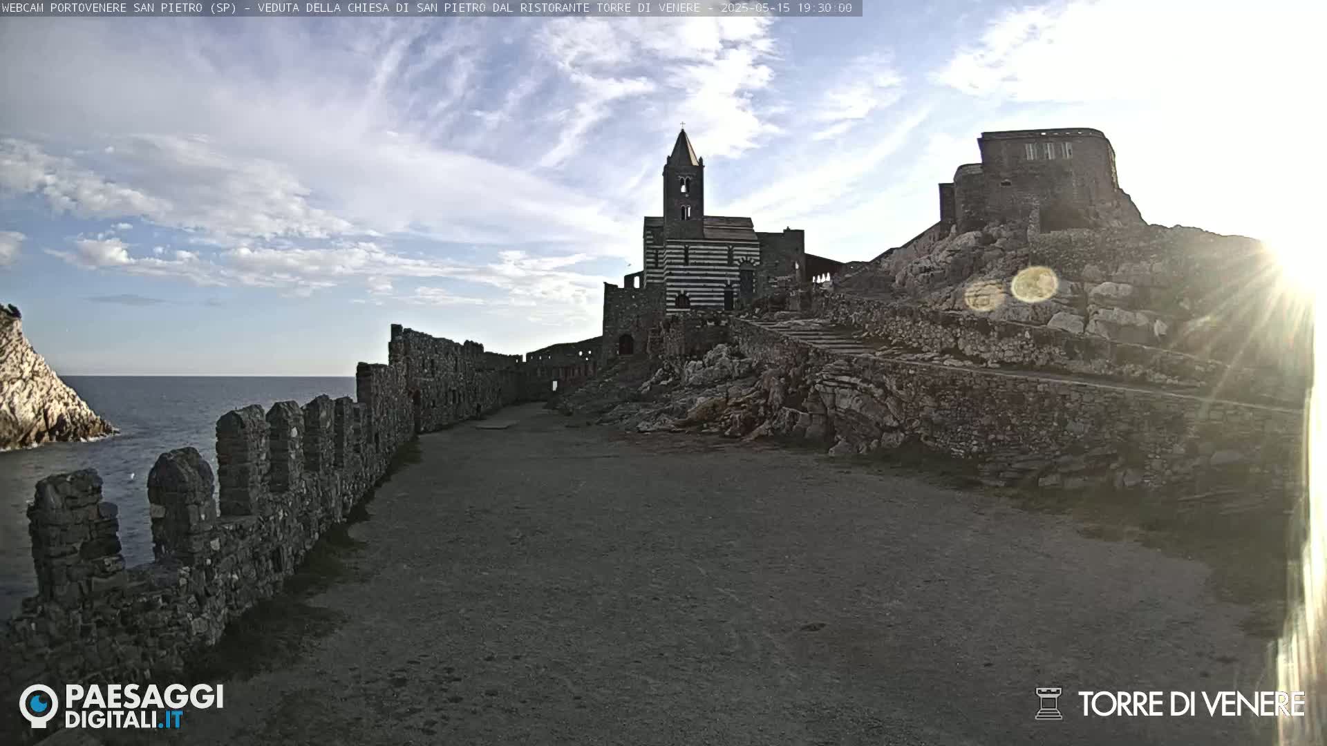 A partly cloudy day reveals a stone pathway leading to a striped church and castle ruins overlooking the ocean, with a stone wall lining the path.