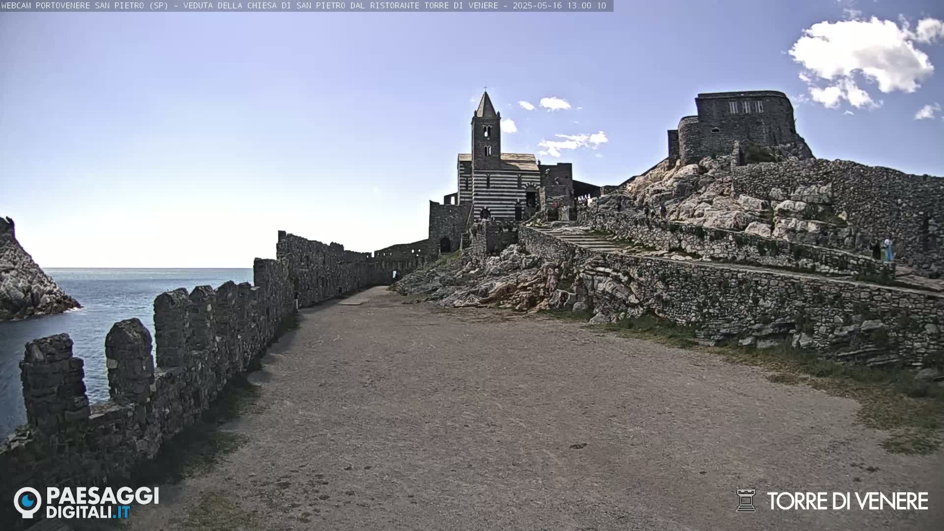 A partly cloudy day reveals a coastal stone church and castle ruins, with a stone pathway leading to them, alongside a low stone wall running near the ocean.