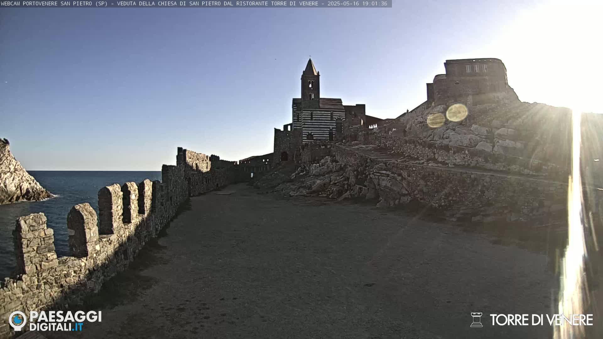 A stone church and castle ruins are situated on a rocky hillside overlooking a calm sea under a clear, sunny sky.