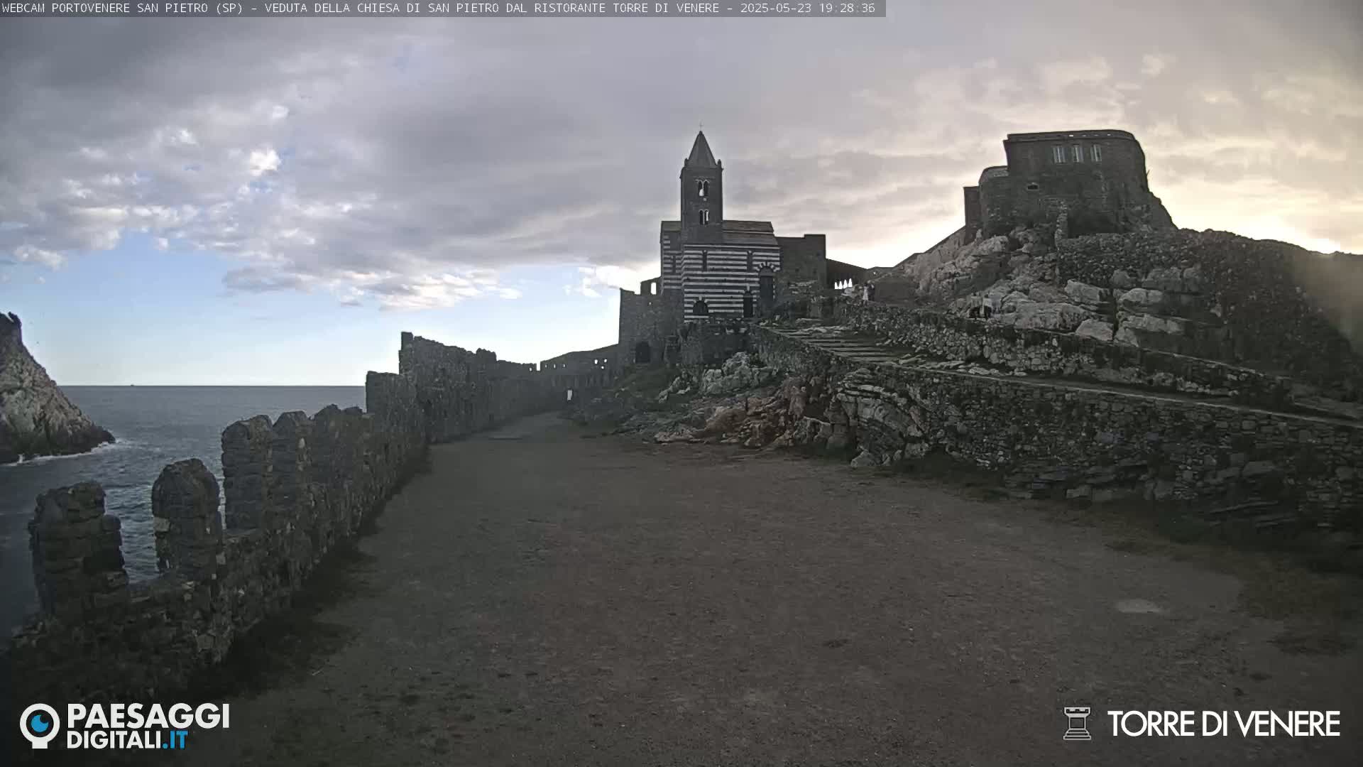 A coastal path winds past ancient stone walls toward a striped church and ruined castle under a partly cloudy sky.