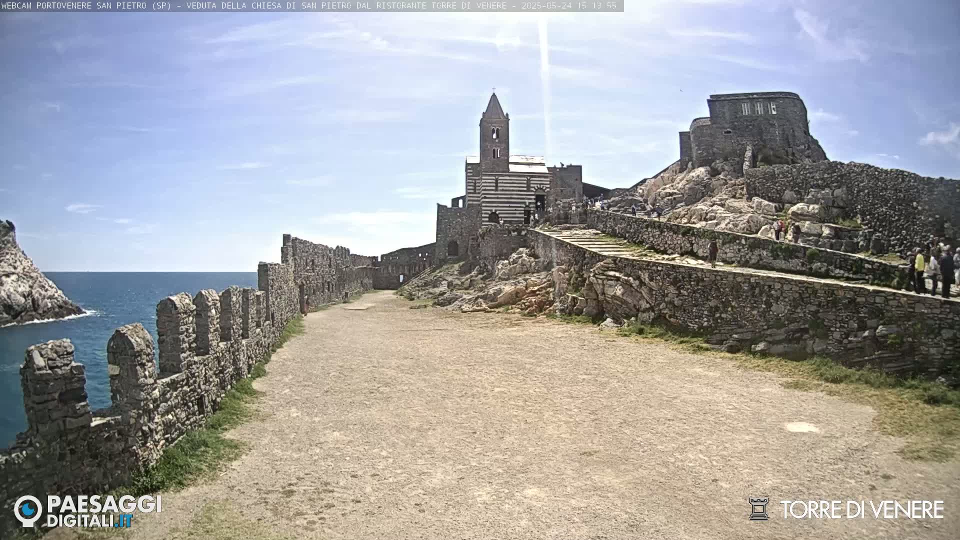 A stone pathway leads to a striped church and castle ruins overlooking a calm ocean under a mostly clear sky.