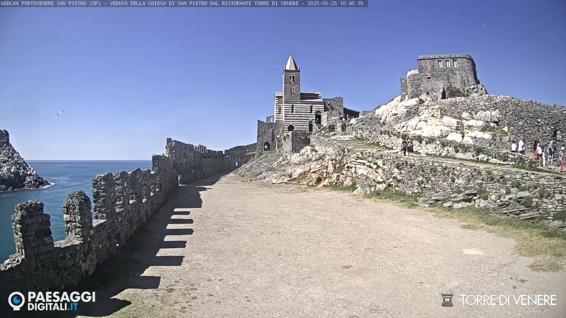 A stone church and castle ruins sit atop a hill overlooking a calm ocean under a clear blue sky.