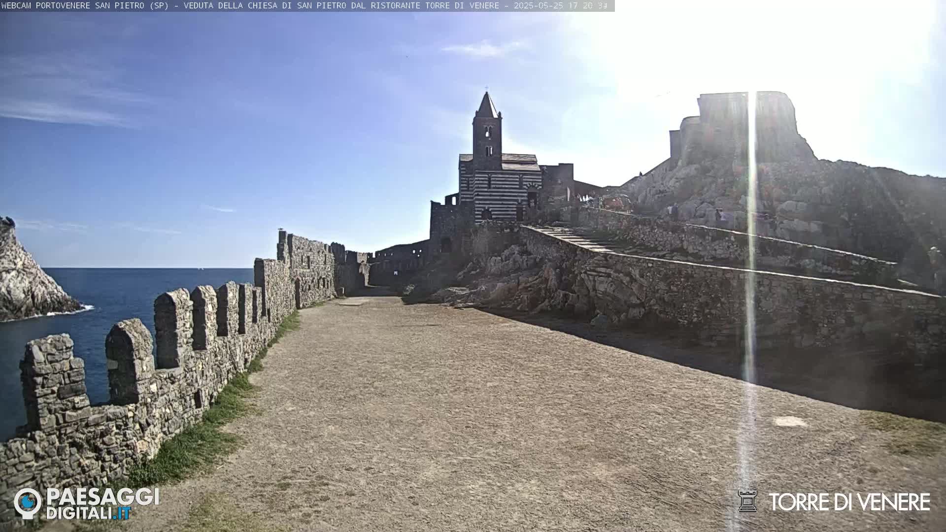 A stone pathway leads toward a striped church and castle ruins overlooking a calm ocean under a bright, sunny sky.