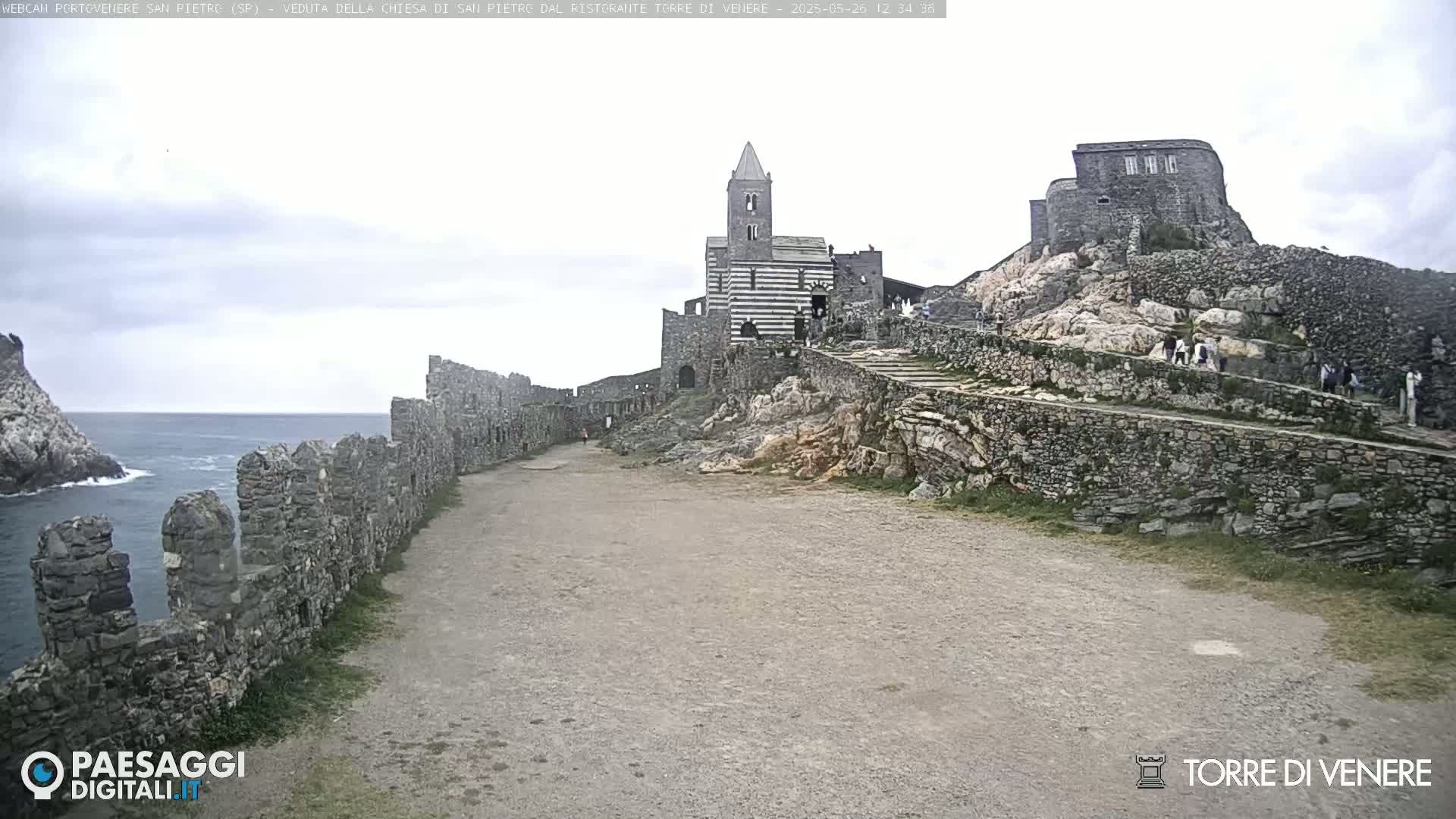 A stone church and castle ruins sit atop a rocky hill overlooking a calm ocean under an overcast sky.