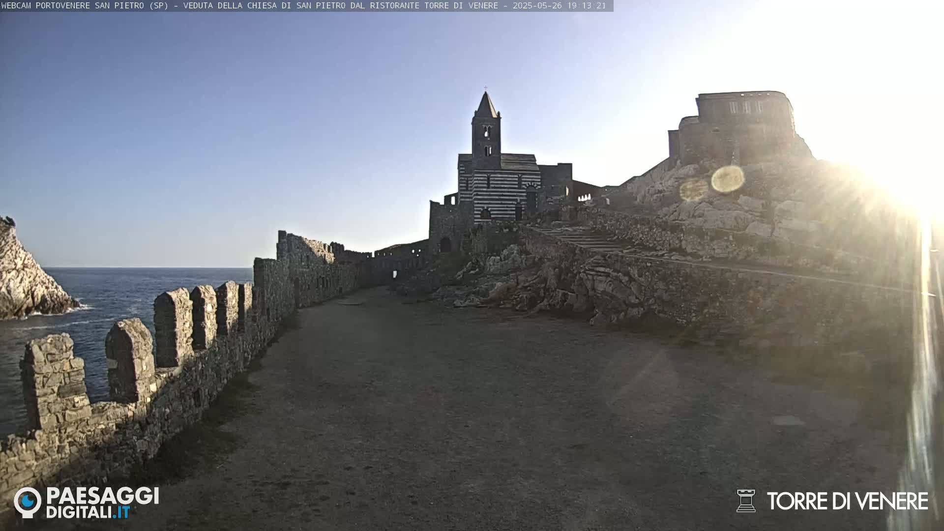 A stone path leads toward a striped church and castle ruins overlooking a calm ocean under a sunny sky.