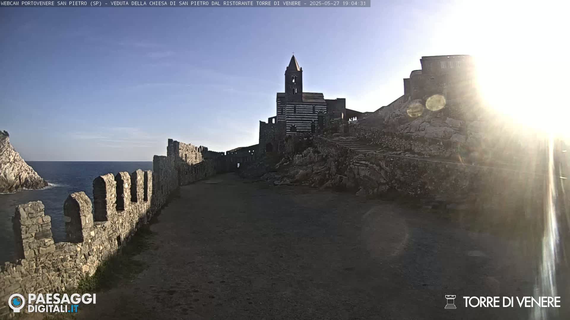 A stone pathway leads toward a striped church and castle ruins, situated on a rocky coastline under a bright, sunny sky.