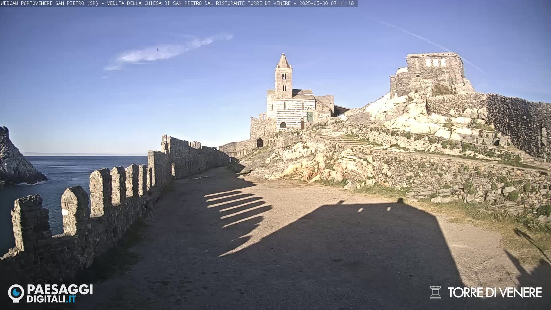 A stone church and castle ruins overlook a calm ocean under a clear blue sky.