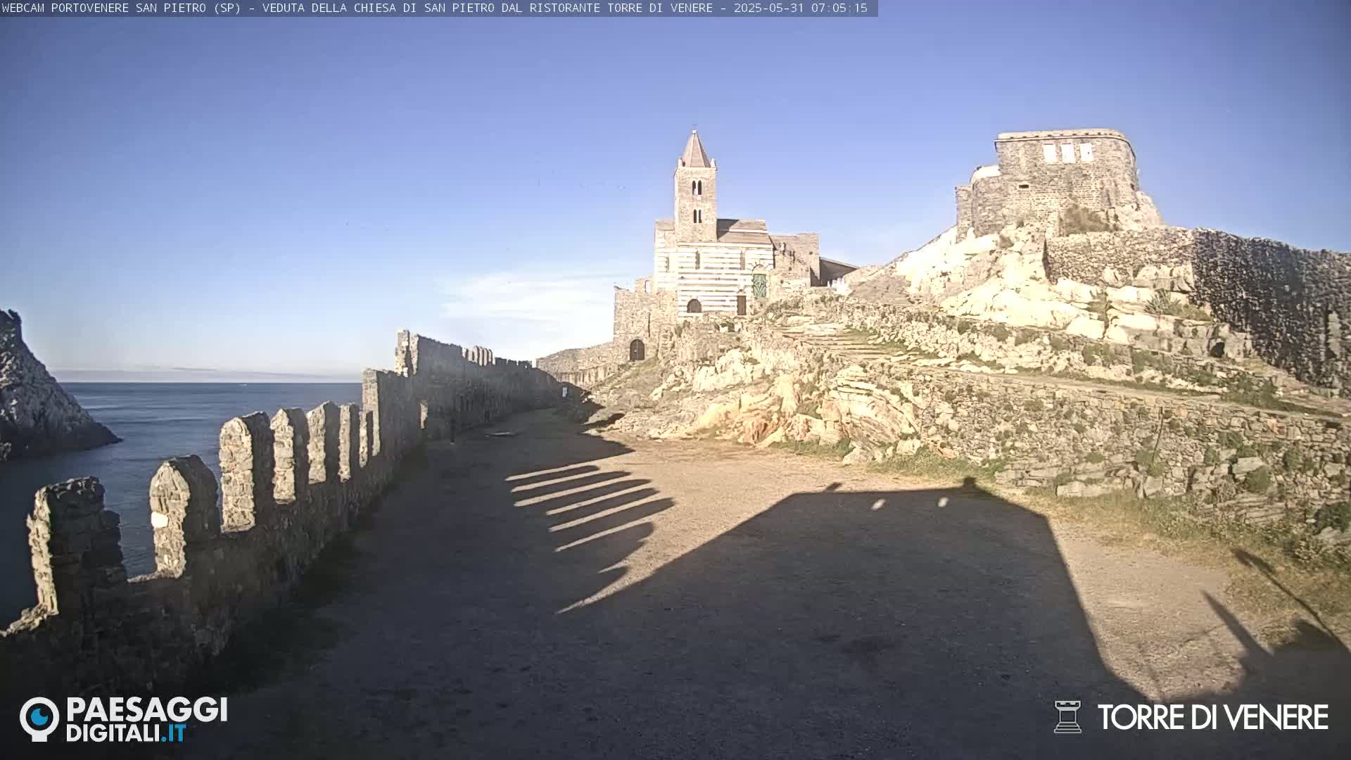 A stone church and castle ruins on a rocky hillside overlooking a calm ocean under a clear blue sky.