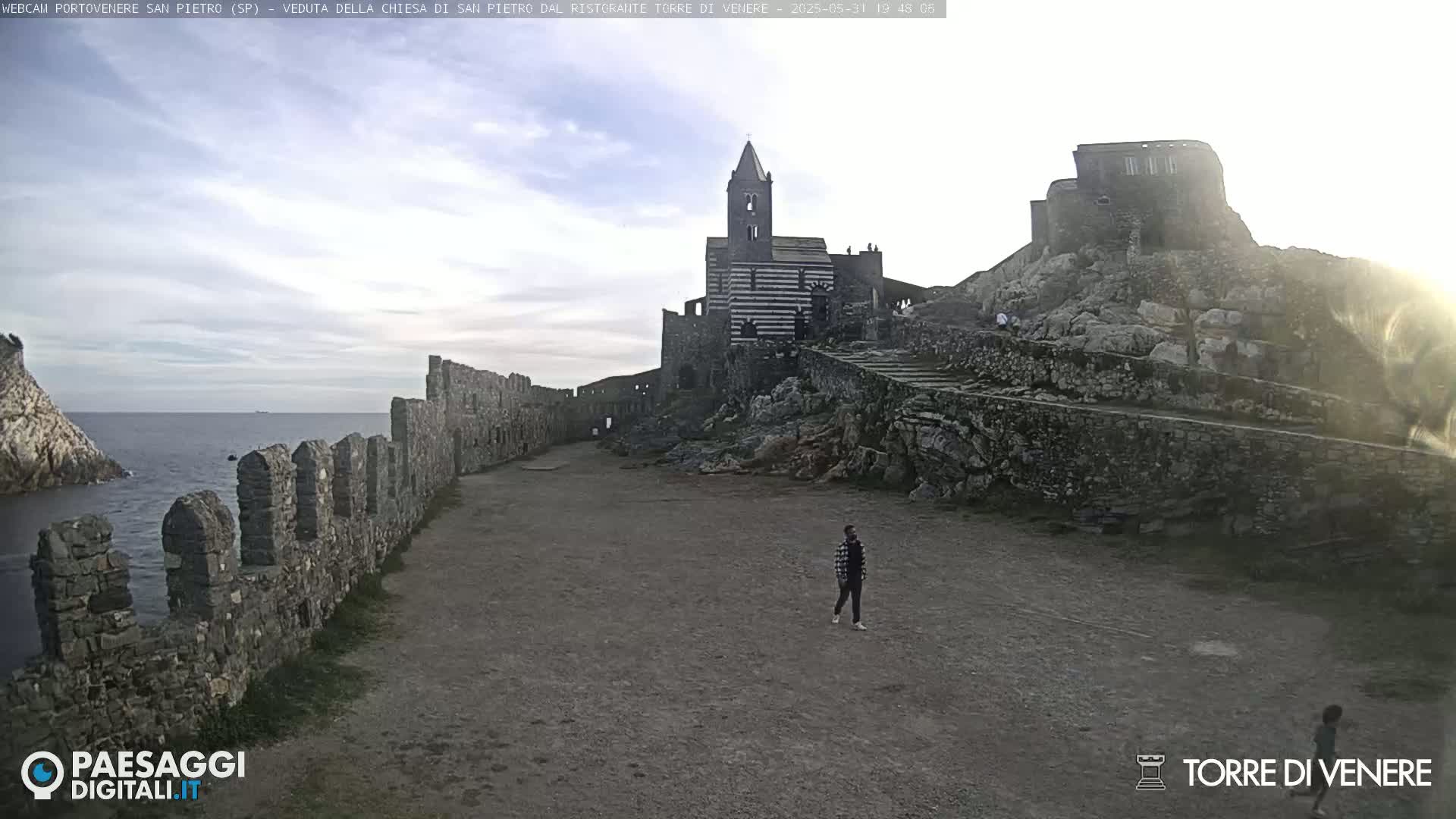 A stone pathway leads to a striped church and castle ruins perched on a rocky outcrop overlooking a calm sea under a partly cloudy sky.