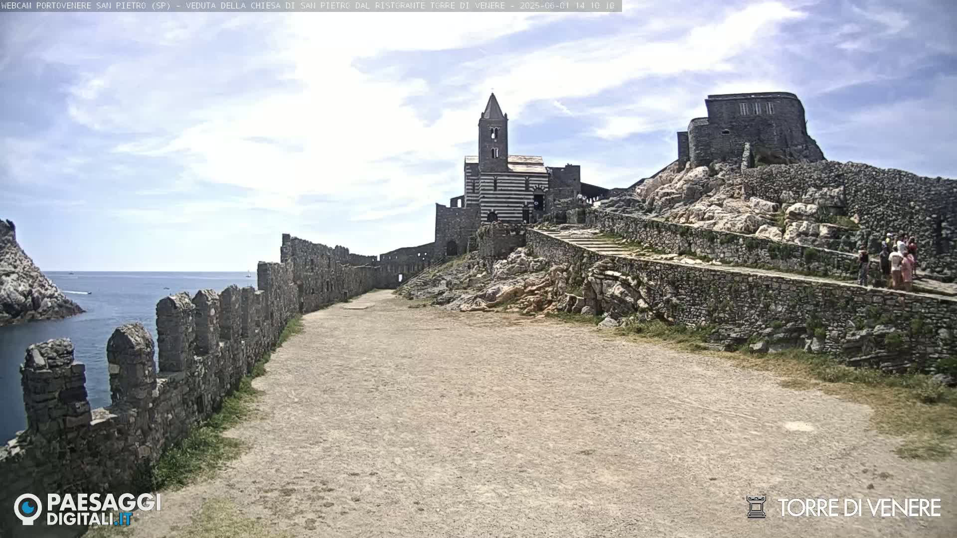 A stone pathway leads toward a striped church and castle ruins situated on a rocky hill overlooking a calm ocean under a partly cloudy sky.
