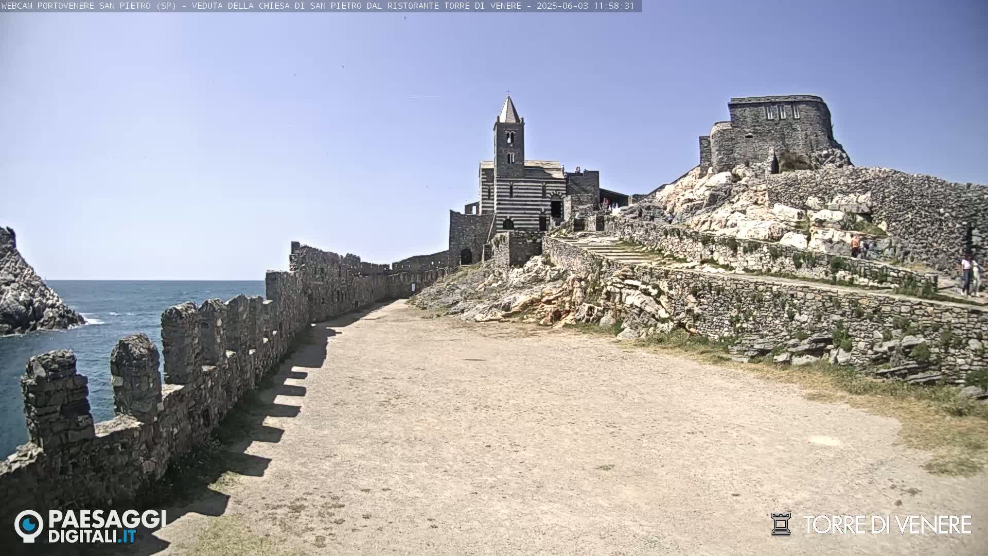 A stone path leads toward a striped stone church and castle ruins perched on a rocky cliff overlooking a calm sea under a clear blue sky.